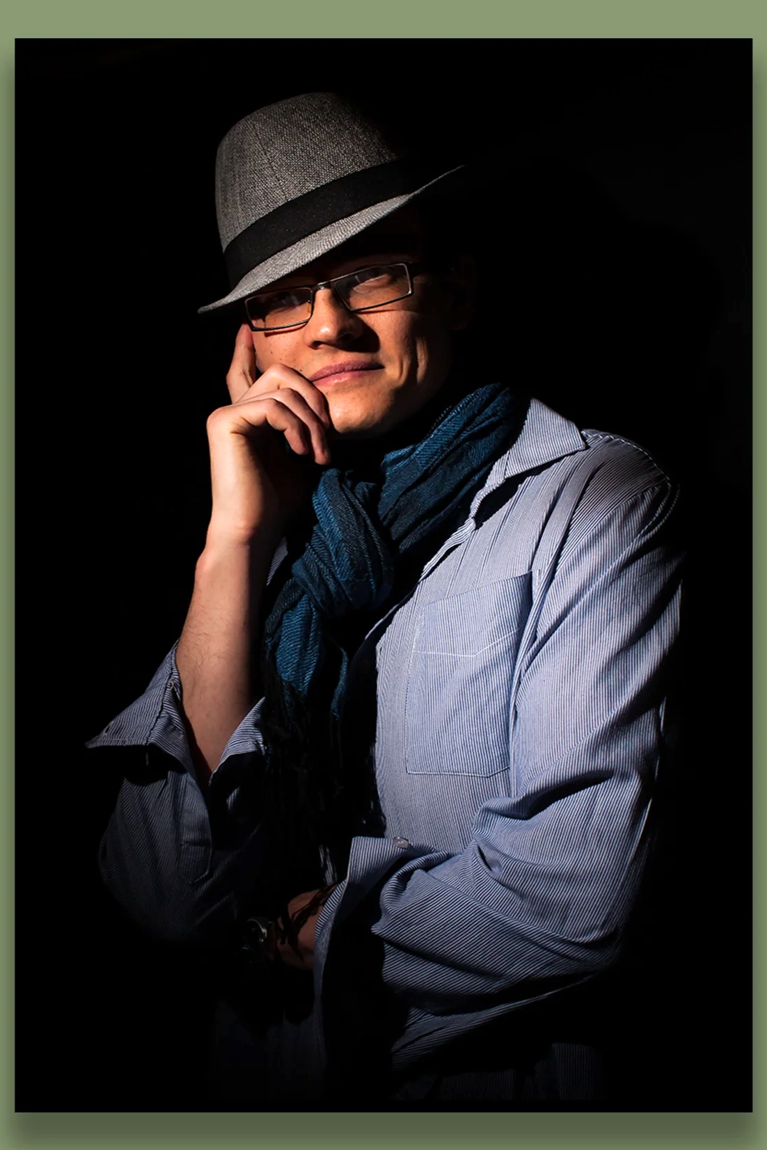 A man wearing glasses, a gray fedora hat with a black band, a blue scarf, and a light gray shirt, posing with one hand near his chin against a dark background.