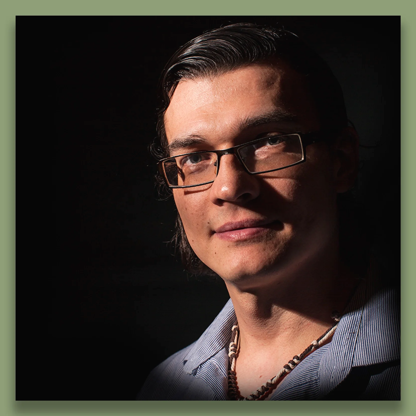 Portrait of a man with dark hair, glasses, and a necklace, wearing a collared shirt, in low light with shadows on his face against a black background.