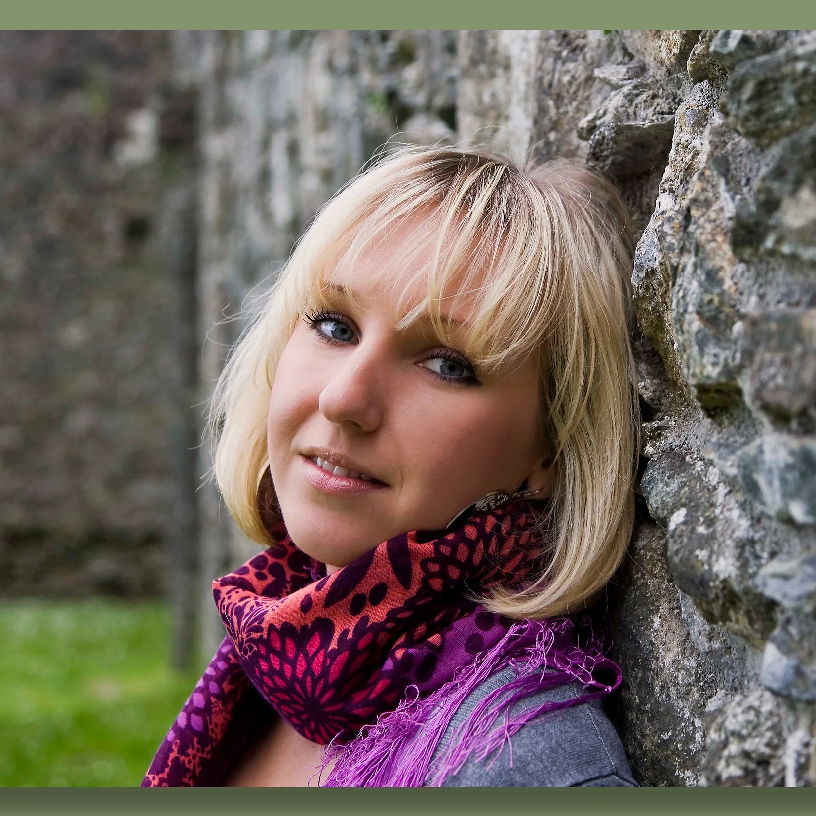 A woman with blonde hair and blue eyes leaning against a stone wall outdoors, wearing a colorful scarf.