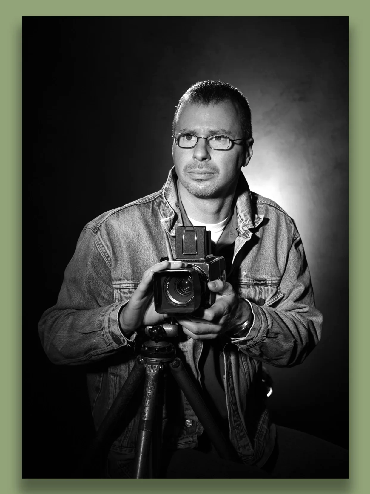 Black and white portrait of a man wearing glasses and a denim jacket, holding a camera on a tripod, looking pensively to the side.