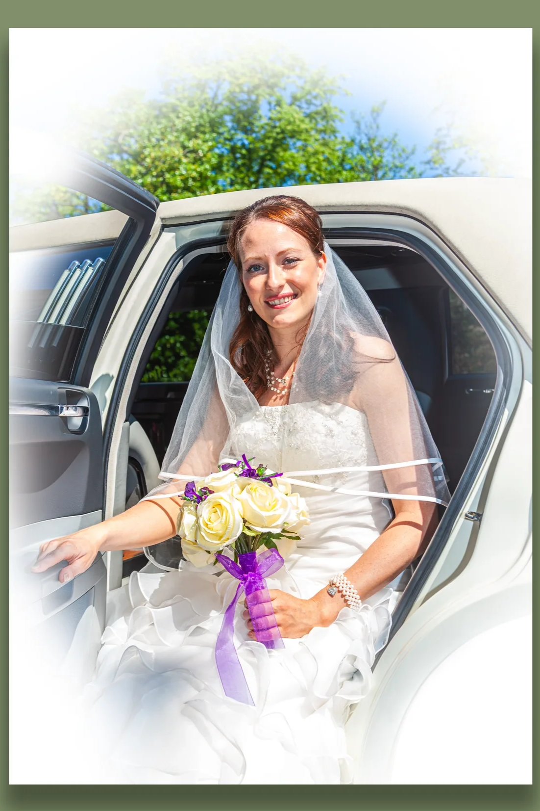 A bride sitting inside a white limousine, smiling and holding a bouquet of white roses and purple flowers, wearing a wedding dress and veil on a sunny day with green trees in the background.