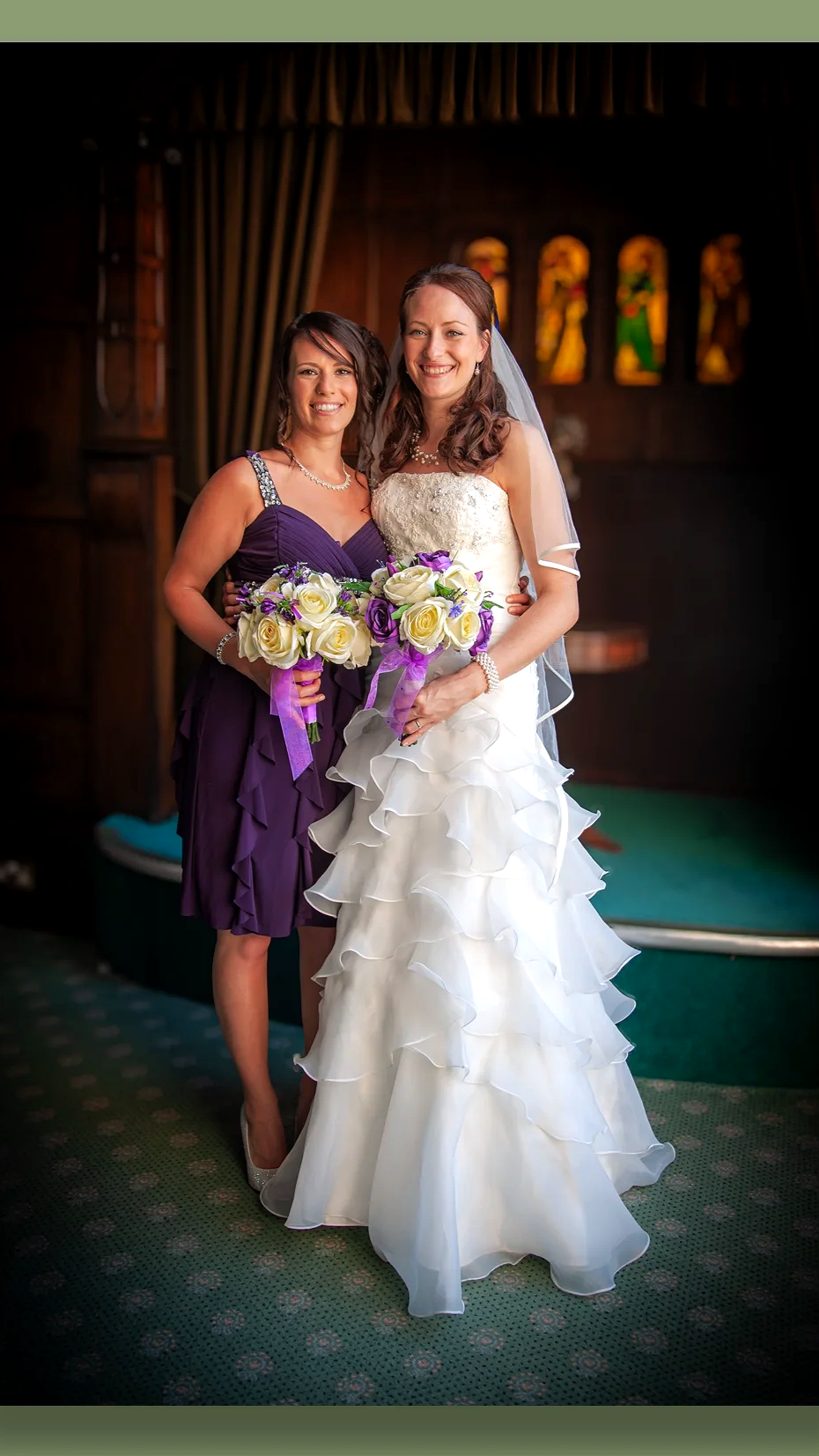 A bride in a white wedding gown holding a bouquet of roses standing next to a bridesmaid in a purple dress holding a similar bouquet inside a church with stained glass windows and dark wood decor.