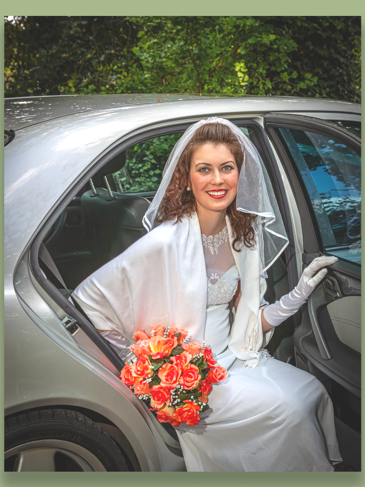 A bride sitting in the back seat of a silver car, wearing a white wedding dress, veil, and gloves, holding a bouquet of pink and orange roses, smiling with greenery as the background.