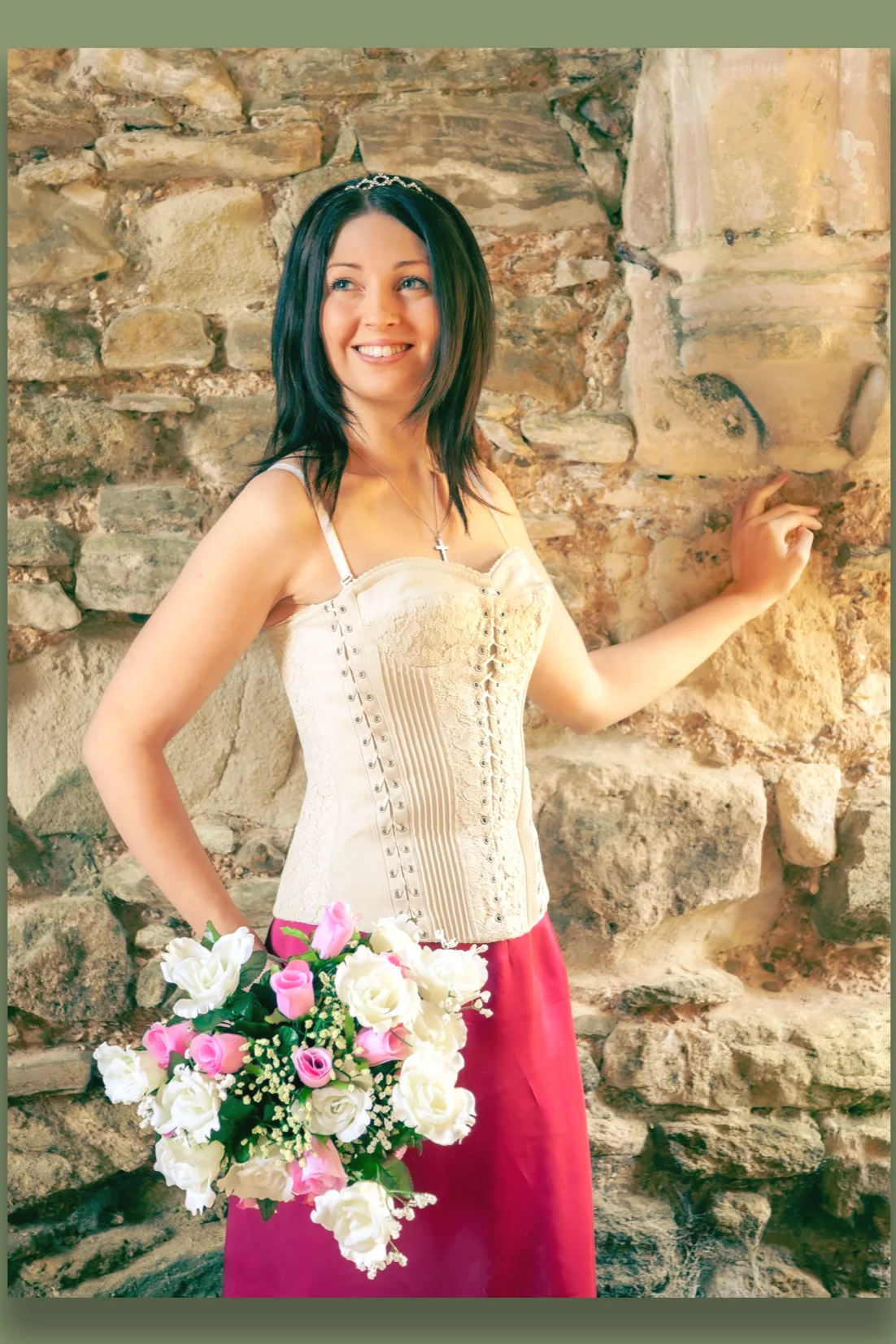 A woman with dark hair wearing a cream-colored corset top and a red skirt, holding a bouquet of pink and white roses, standing against a stone wall, smiling and looking to the side.