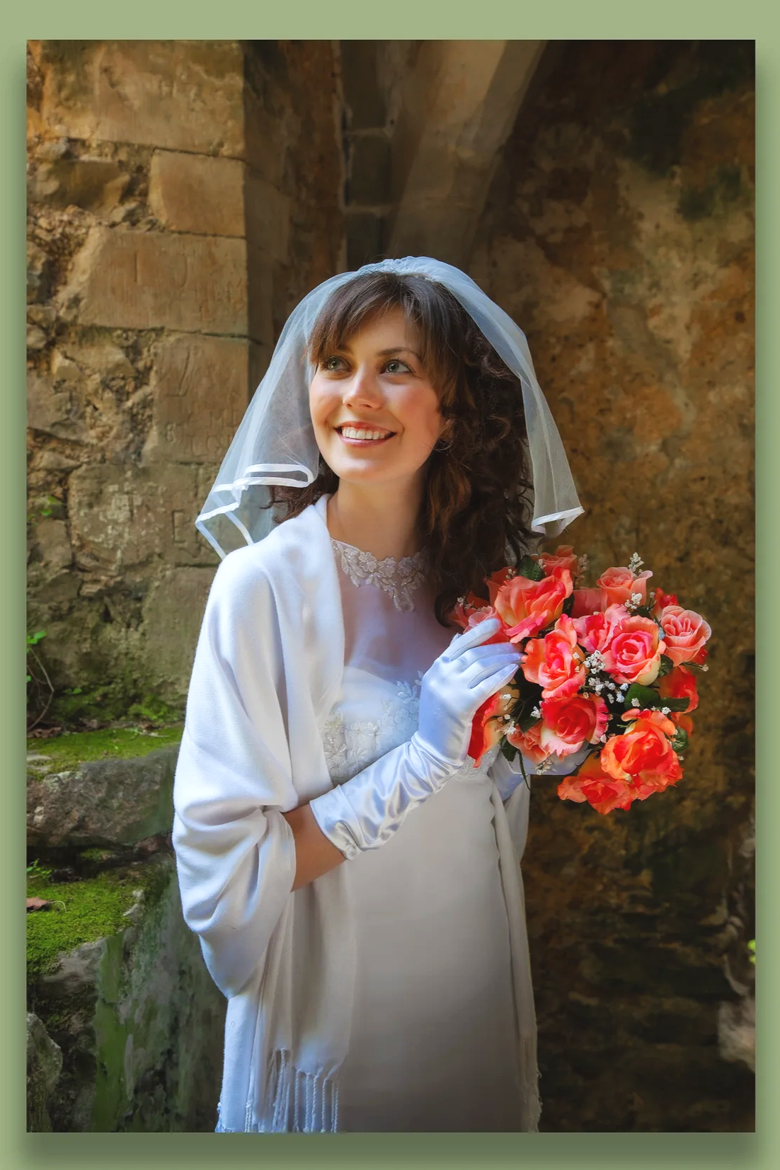 A young woman in a white wedding dress and veil holding a bouquet of pink and orange roses, standing outdoors near an old stone wall, smiling and looking to her left.