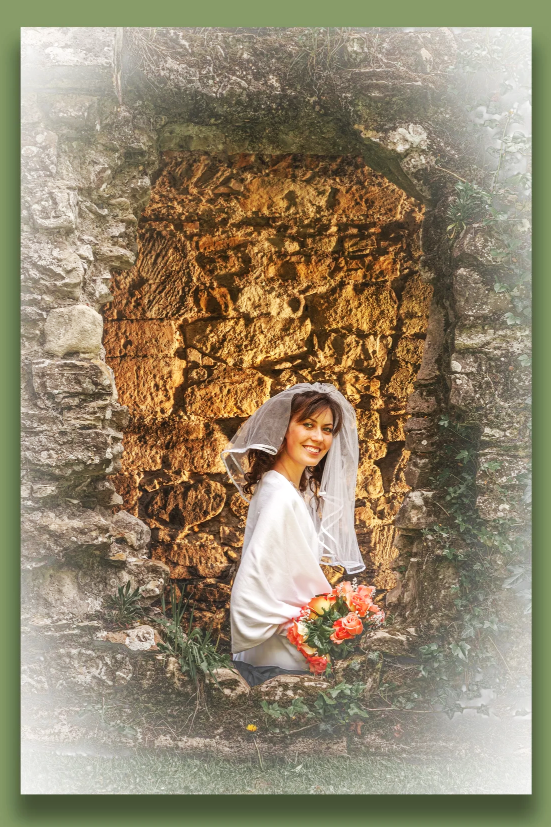 A young bride,  wearing a white blouse and a veil, holding a bouquet of pink and orange flowers, and smiling, standing in front of an old stone wall with a rounded archway.