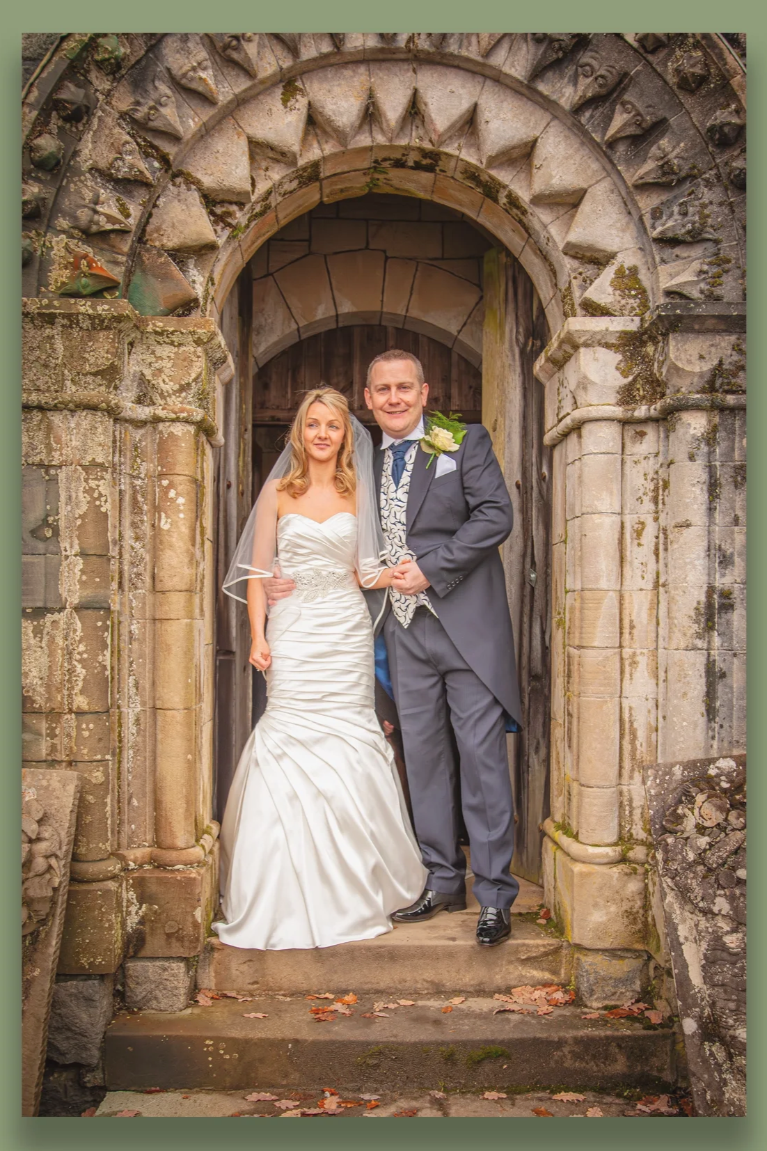 A bride and groom standing in front of a historical stone archway, dressed in wedding attire, smiling for the camera.