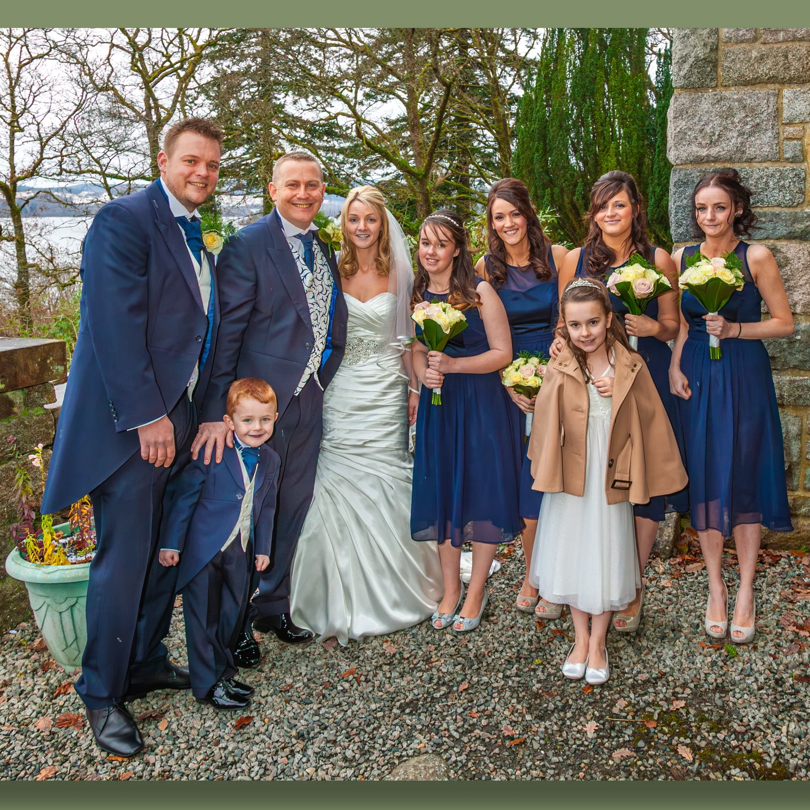 Group of people at a wedding, including the bride in a white gown and veil, the groom, and eight bridesmaids and children, standing outdoors on a gravel path near trees and a stone wall.