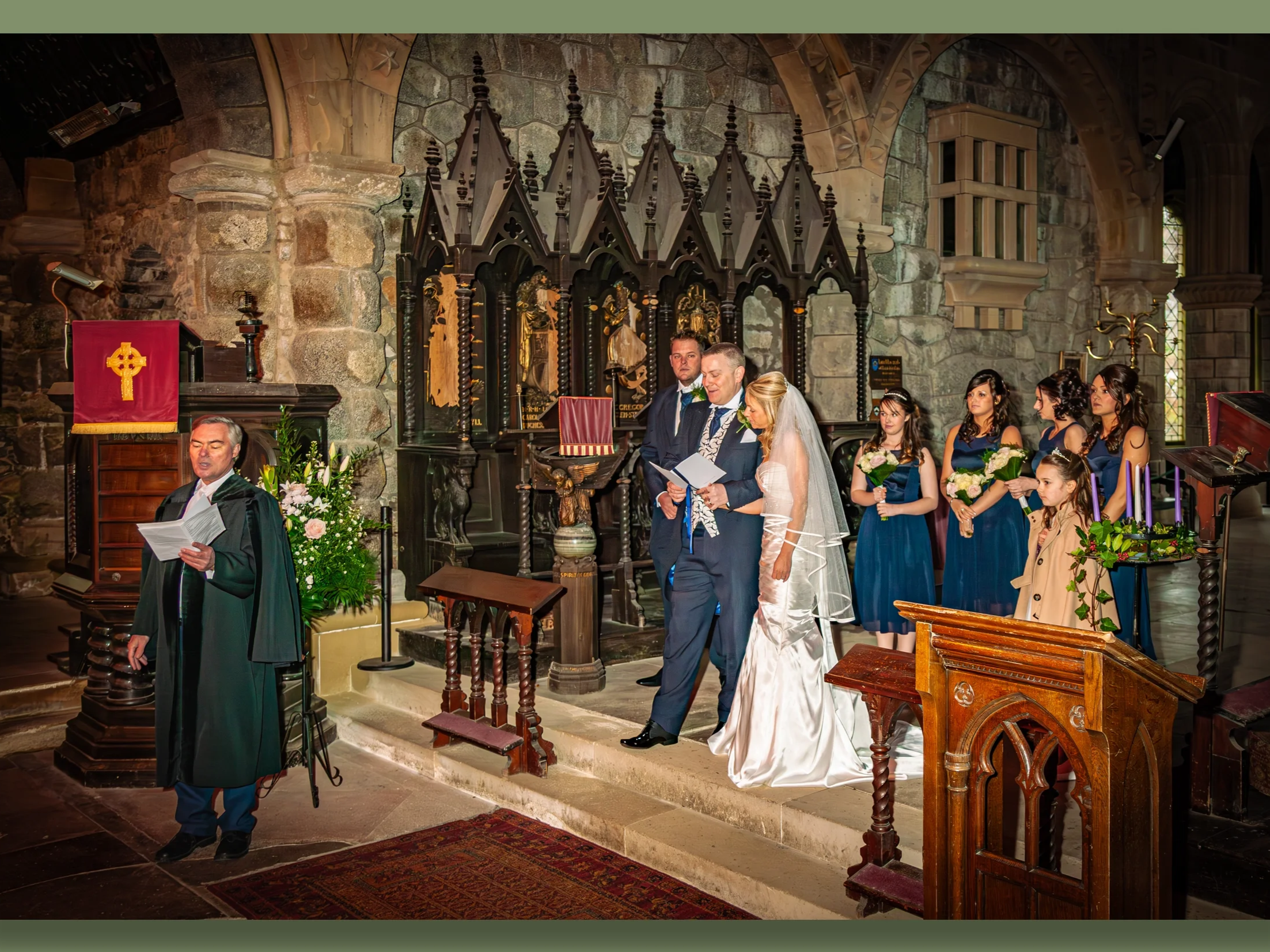 A wedding ceremony inside a stone church with a priest, bride, groom, and bridesmaids dressed in blue, with the bride in a white gown and veil standing beside the groom, who is in a dark suit. The officiant is reading from a book, and there are flora