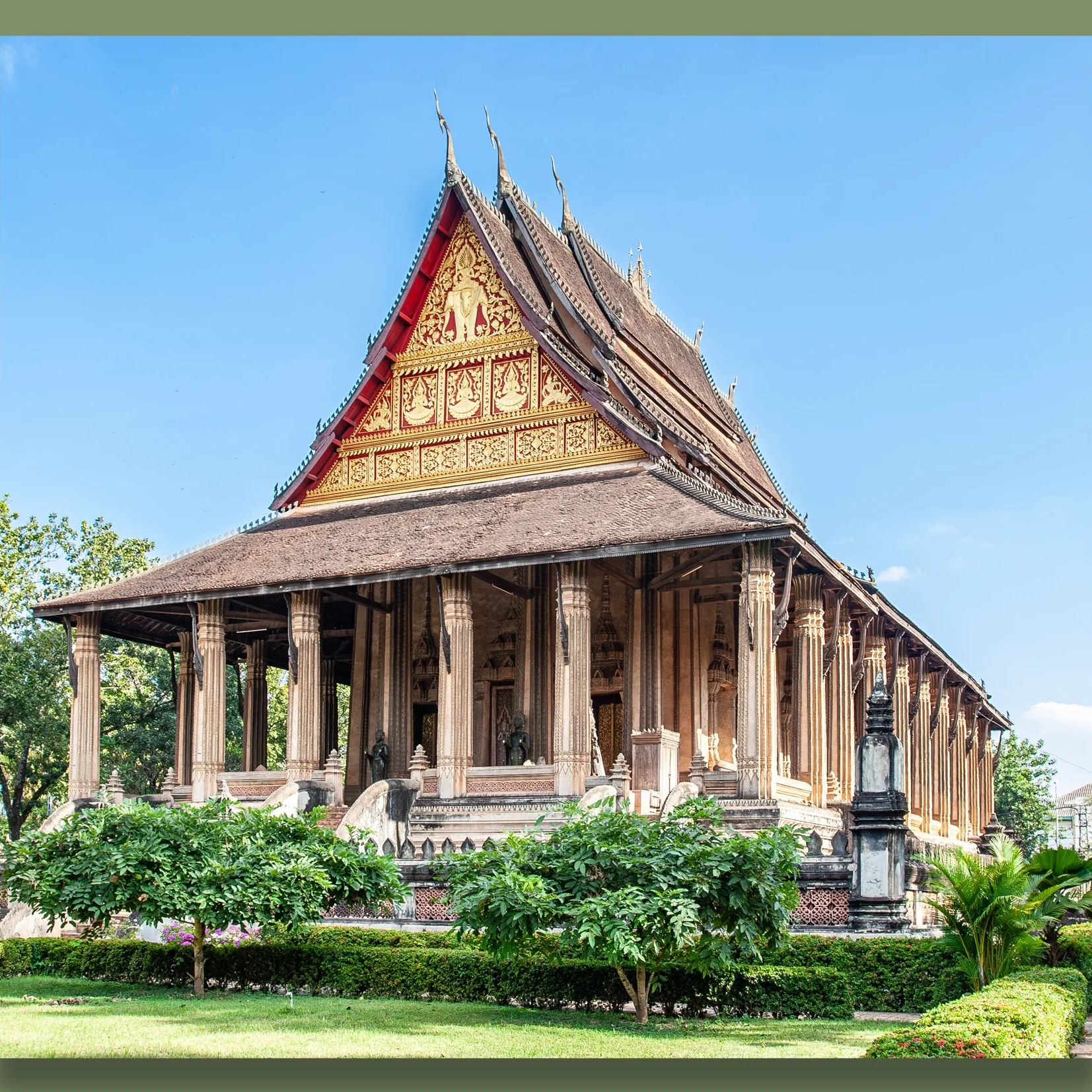A traditional temple in Vientiane, with ornate gold and red decorations, wooden pillars, and a steep, multi-tiered roof, surrounded by green trees and shrubs under a blue sky.