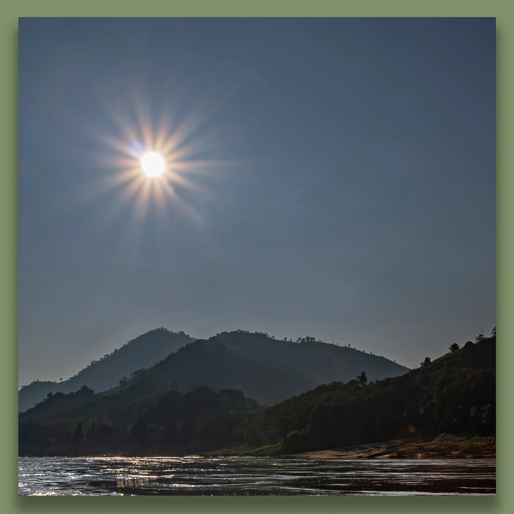 The sun shining brightly in a clear sky over mountain hills and the Mekong river in the foreground.