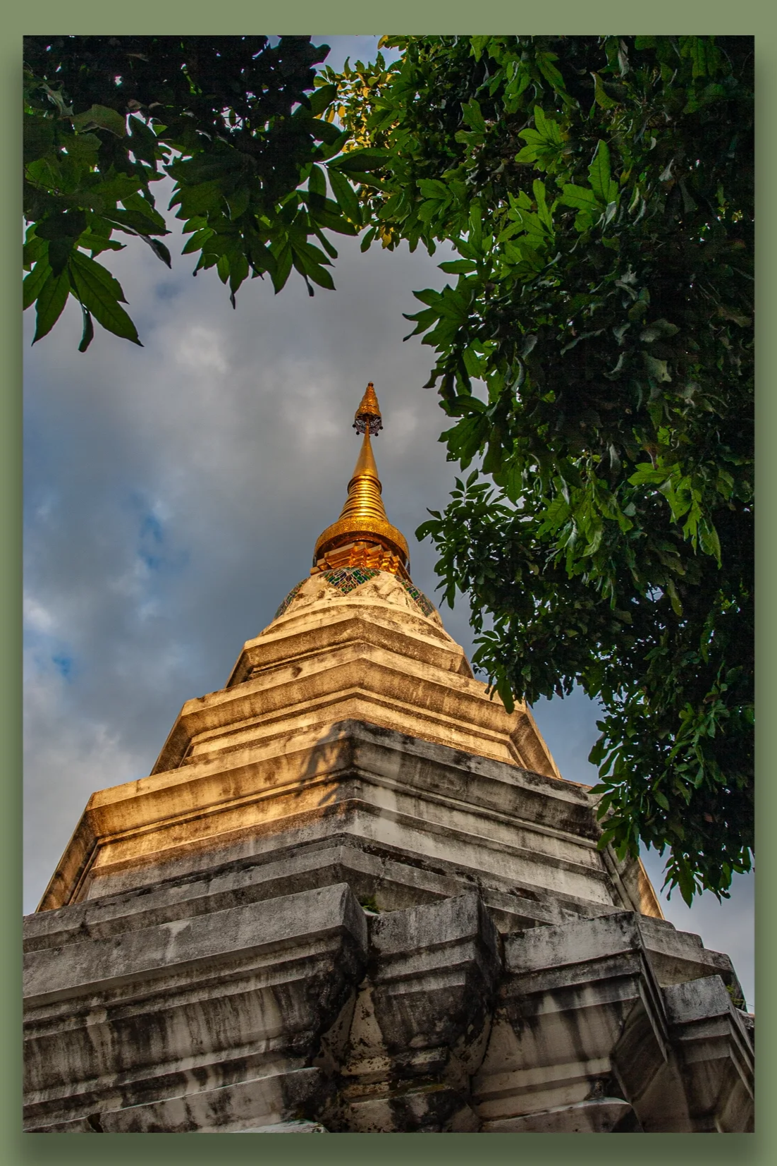 A tall, ancient stone and gold temple spire partially surrounded by green tree leaves, against a cloudy sky.