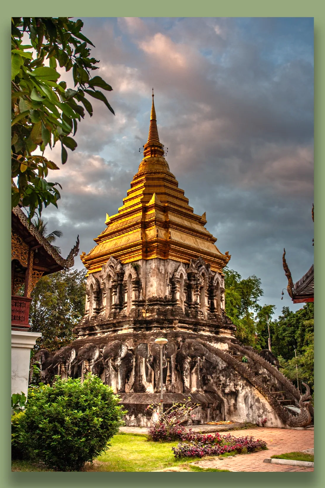 A historic Buddhist temple with a golden stupa, surrounded by greenery and traditional-style buildings, set against a cloudy sky at sunset.