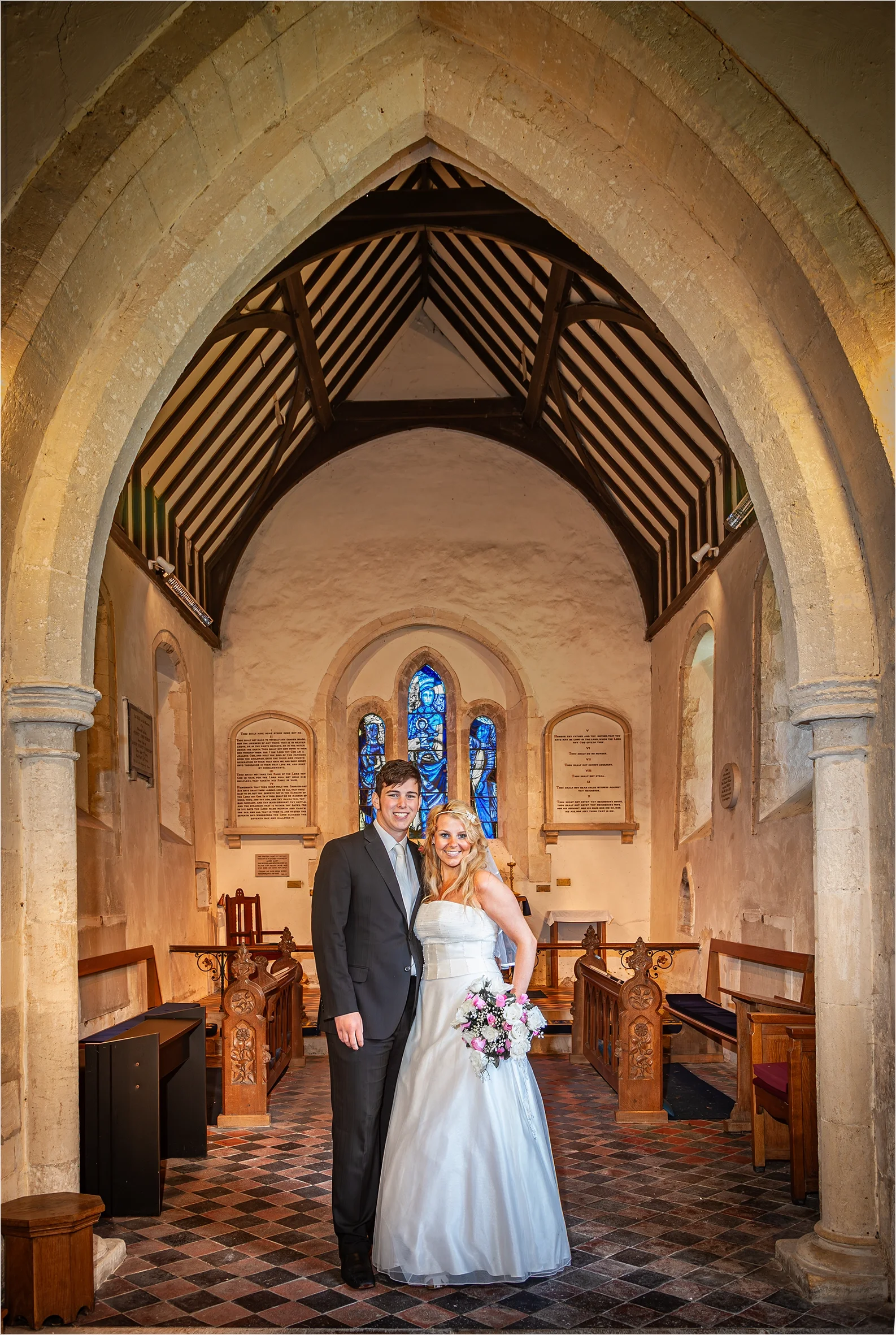 A bride and groom inside a small church, standing close together and smiling, with stained glass windows in the background.