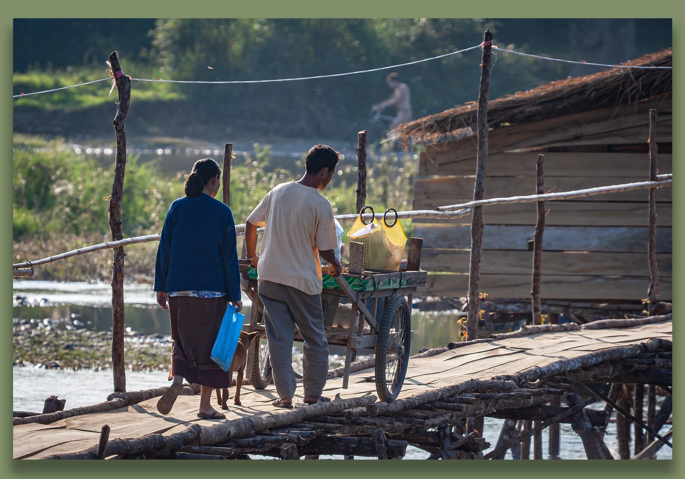 Vang Vieng, Laos.  Two people walking across a wooden bridge over water, one pushing a cart with yellow bags, and a dog walking beside them, with a blurred figure of a person fishing in the background.