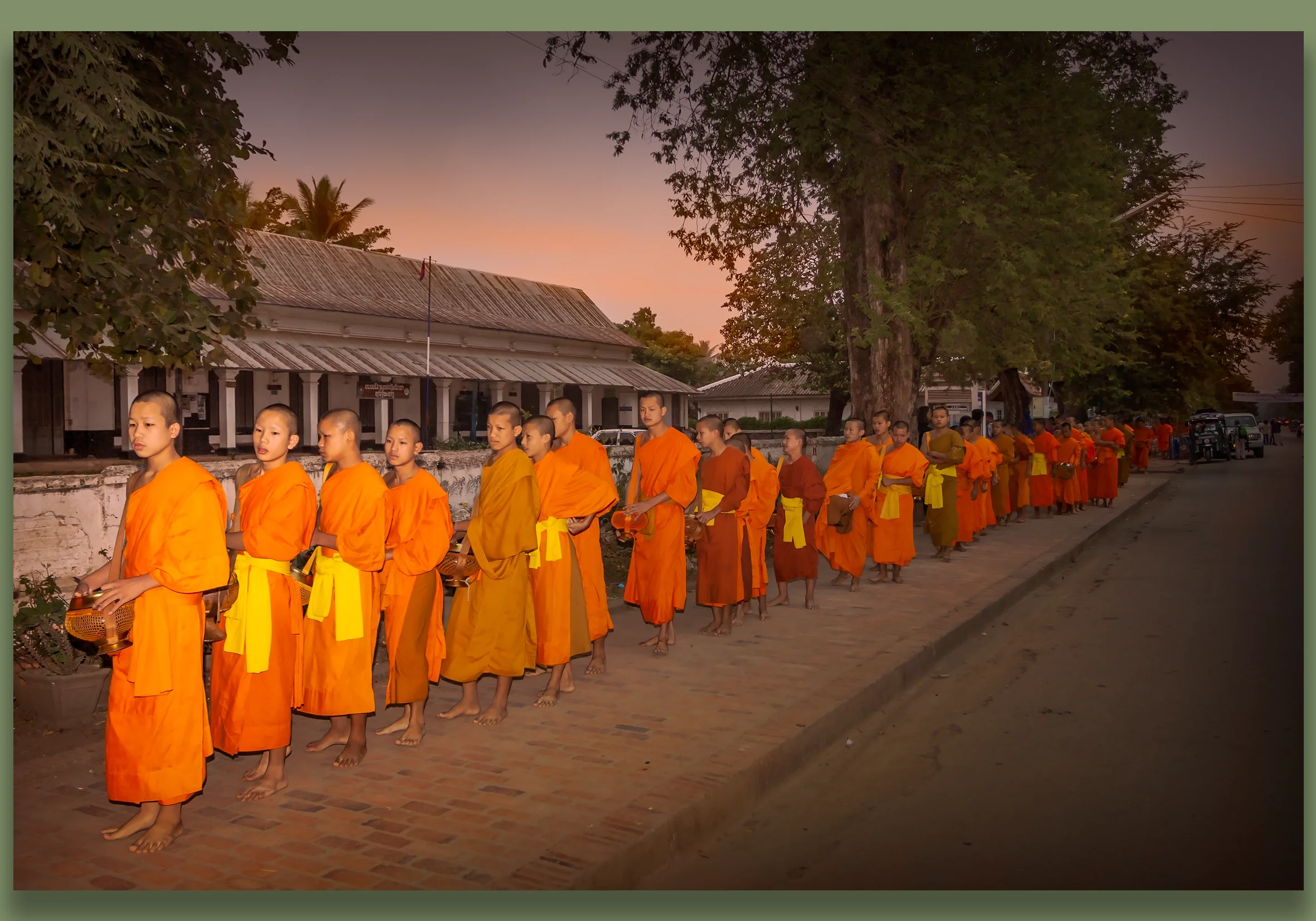 Luang Prabang at sunrise, Buddhist monks walking in a line , dressed in orange robes, some holding alms bowls, on a street  with trees and traditional buildings in the background.