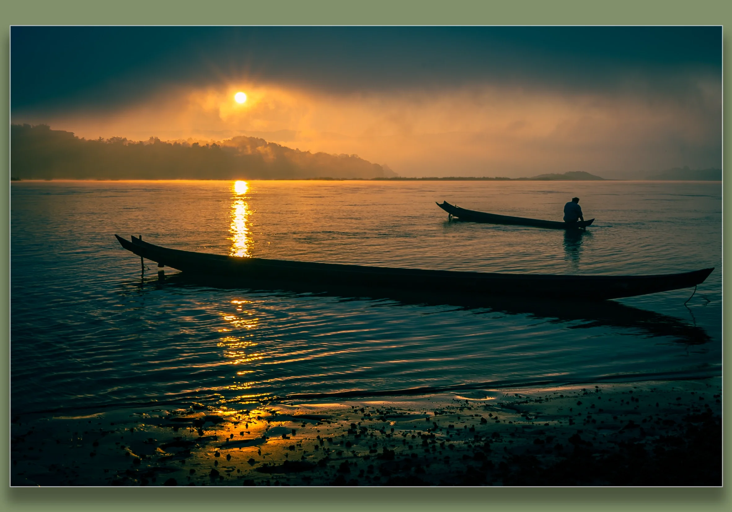Silhouettes of two boats on the Mekong River during sunrise, with one person sitting in one boat, and the sun casting a reflection on the water, with morning mist over the distant shoreline.