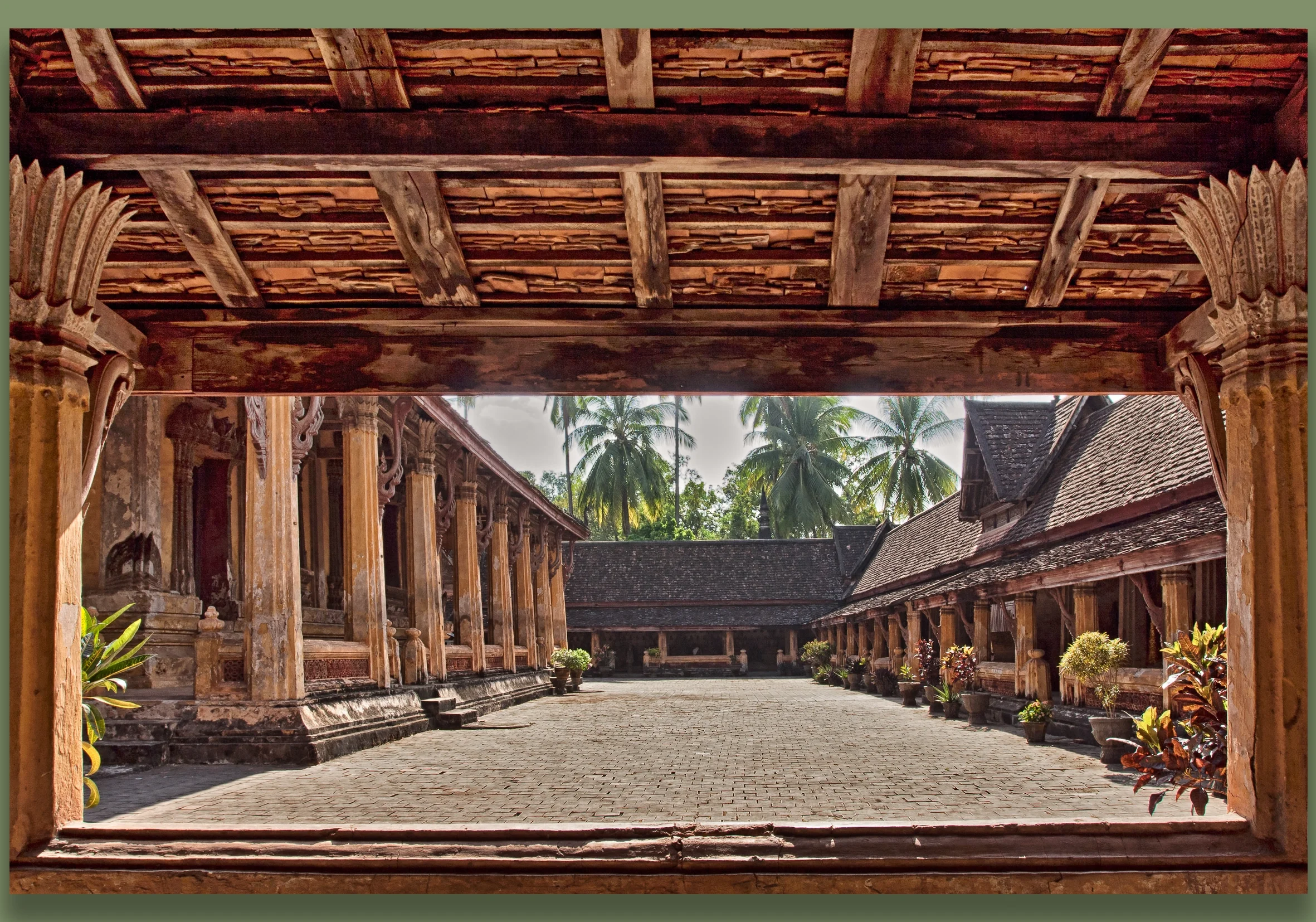 View of an old traditional temple courtyard surrounded by wooden buildings with detailed carvings, and potted plants along the walkway, with palm trees in the background.