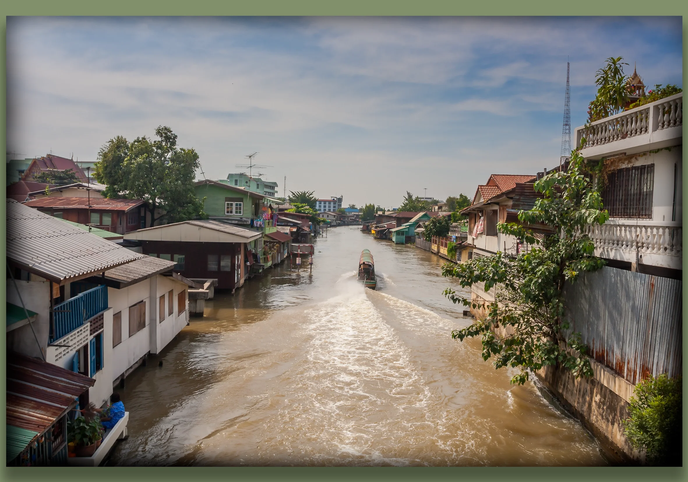 A canal in a residential area of Bangkok, with houses lining both sides and a boat moving through the water, creating ripples. The sky is partly cloudy, and the scene shows a tropical climate with some greenery.