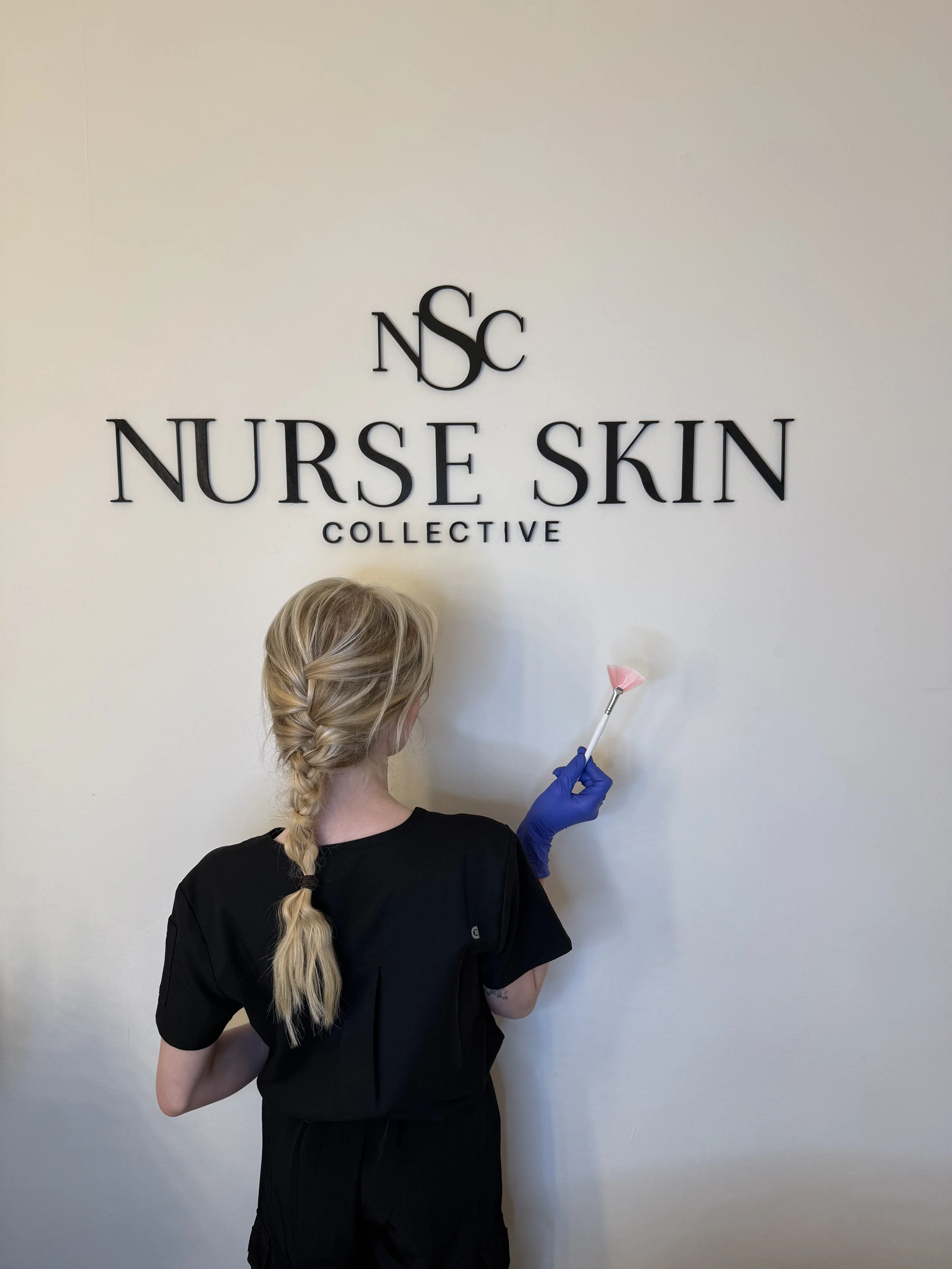 A woman with blonde hair in a braid, wearing a black uniform and purple gloves, cleaning a wall with a pink duster in front of a sign that reads 'Nurse Skin Collective'.