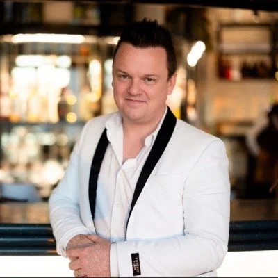 A man in a white blazer and dress shirt with a black camera strap around his neck, standing in a warmly lit indoor setting.