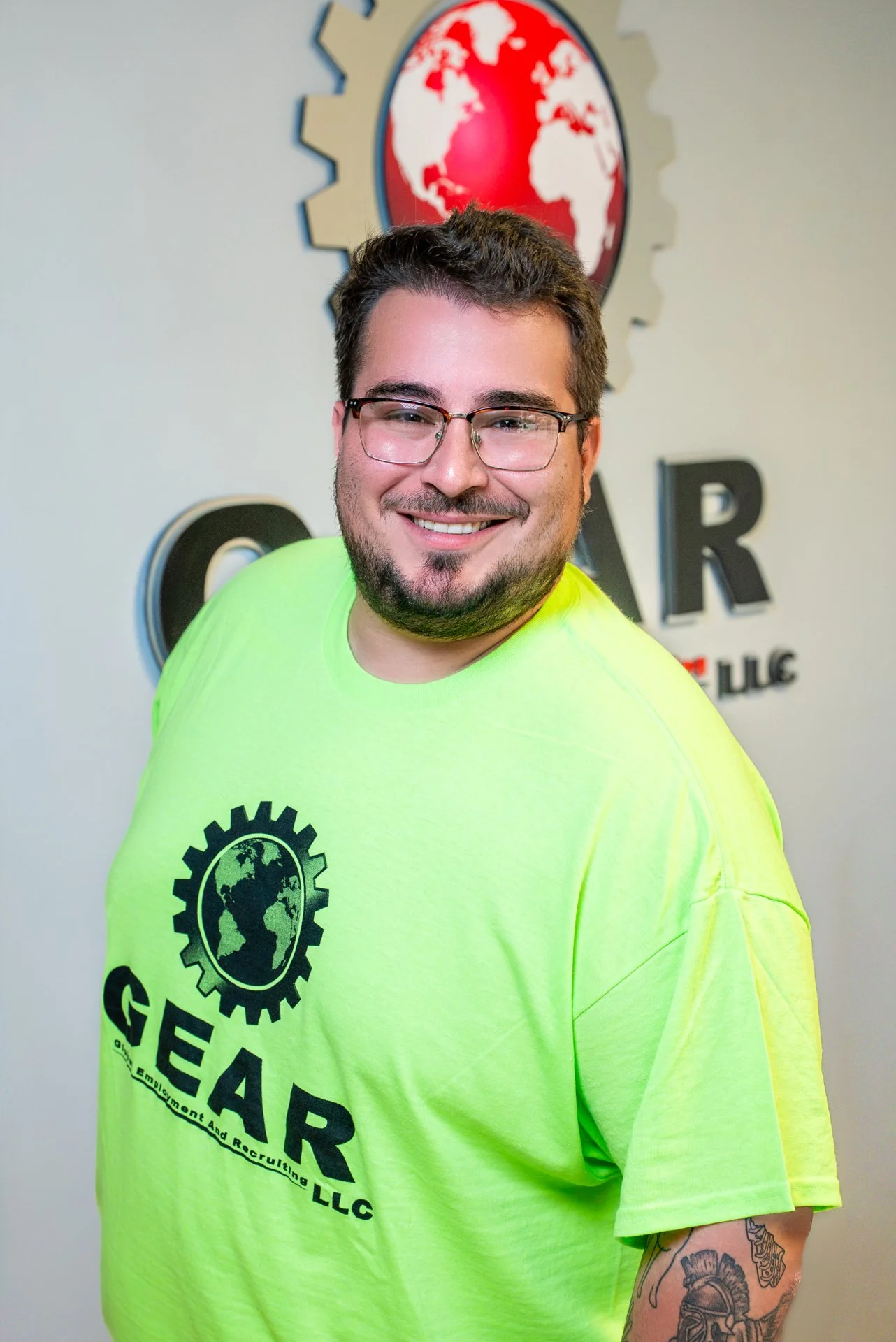 A smiling man with glasses and a beard wearing a bright yellow shirt with the logo "GEAR" and a globe with gears around it, standing in front of a wall with a logo of a red globe inside a gear.