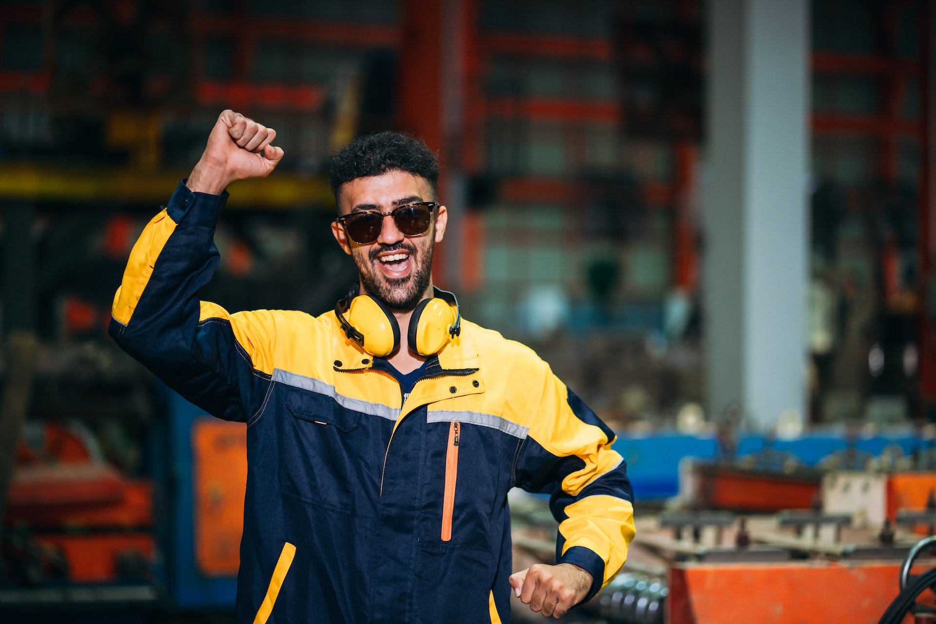 A man smiling and celebrating with a raised fist in an industrial workplace. He is wearing sunglasses, a yellow and blue jacket, with yellow headphones around his neck, in a workshop or factory setting.
