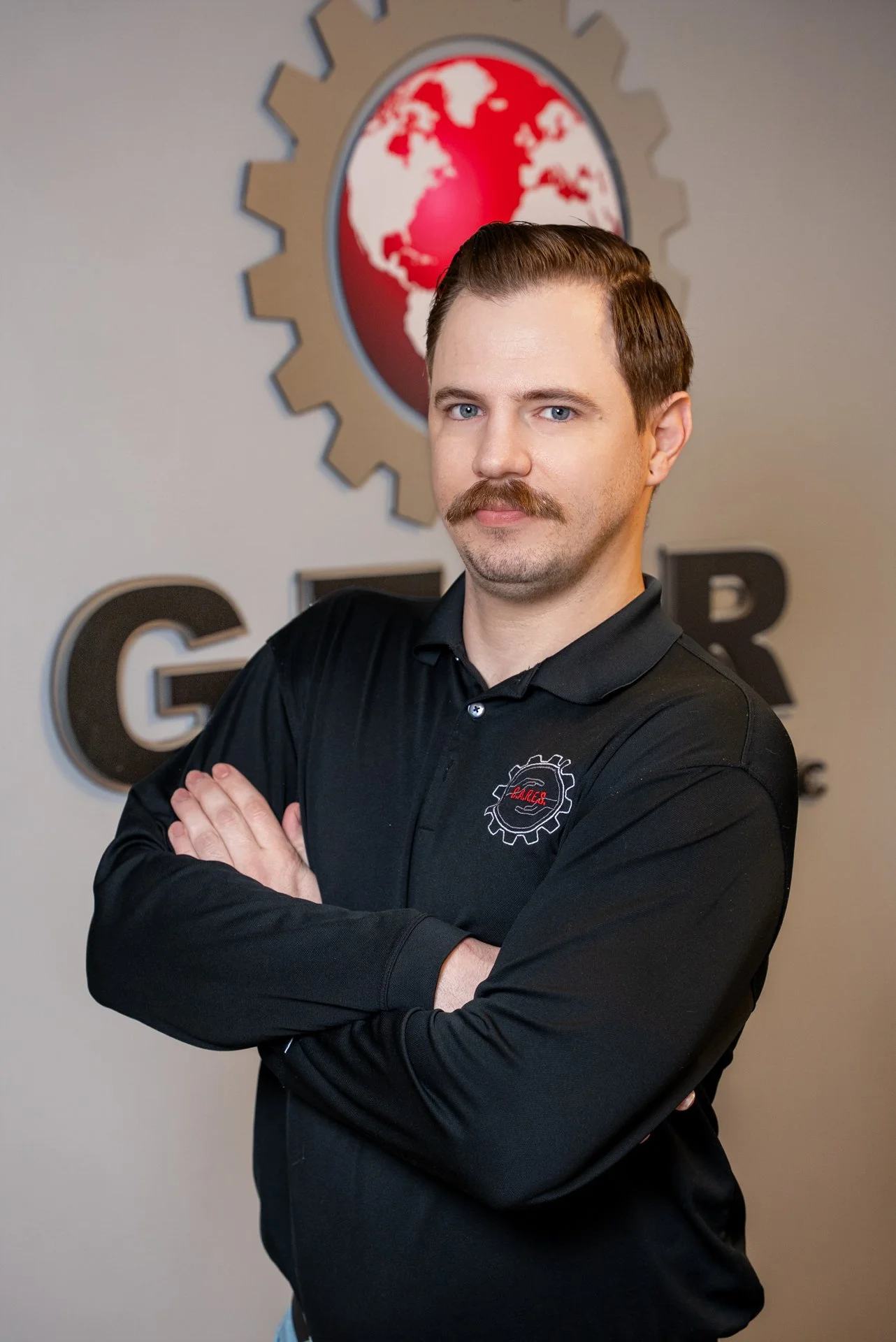 A man with a mustache and short brown hair standing with arms crossed in front of a wall with a logo of a globe surrounded by a gear and the letters GEAR.