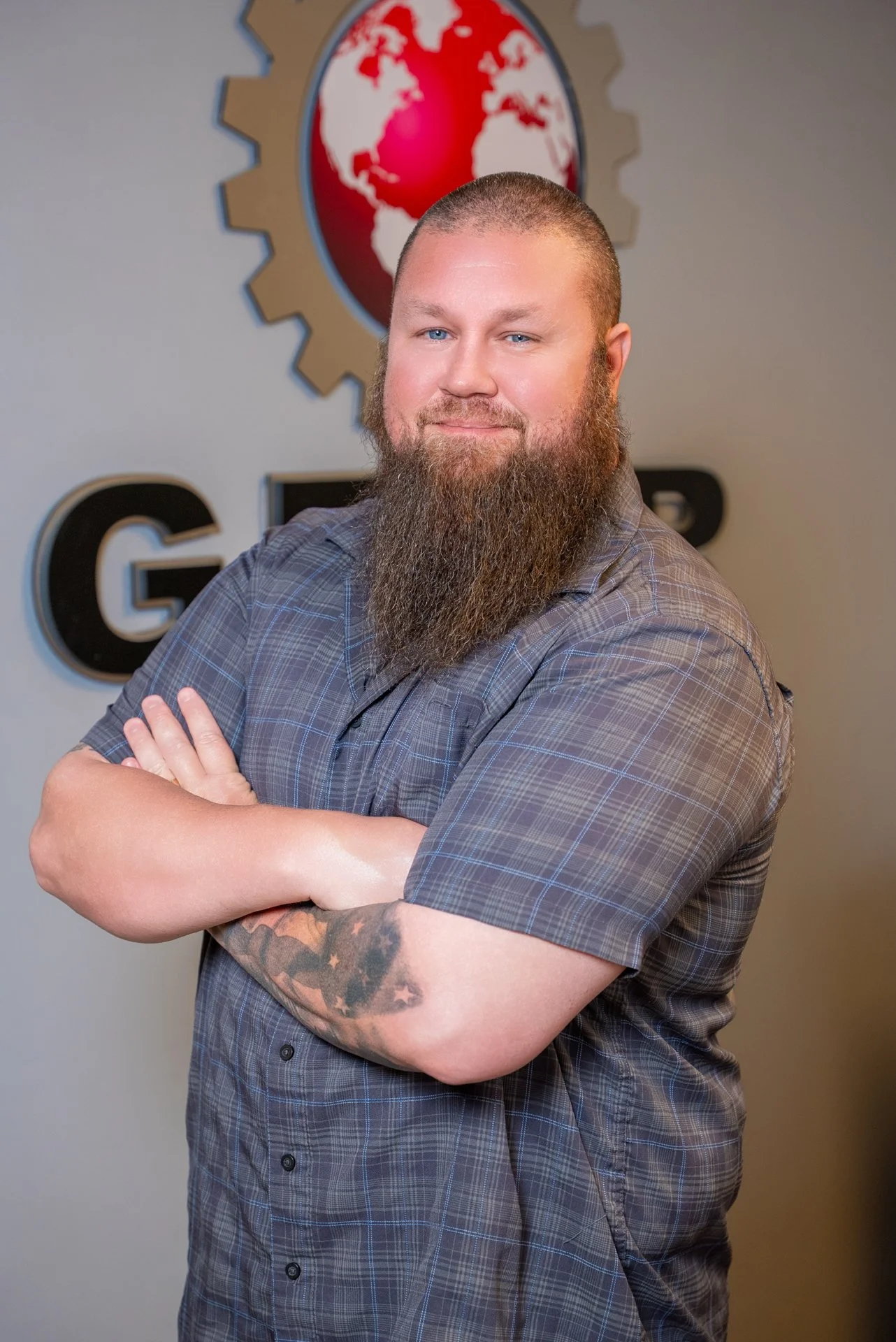 A man with a beard and tattoos on his left arm, wearing a plaid shirt, standing with arms crossed in front of a logo that features a red globe surrounded by a gear, with the partial text 'G...' visible behind him.