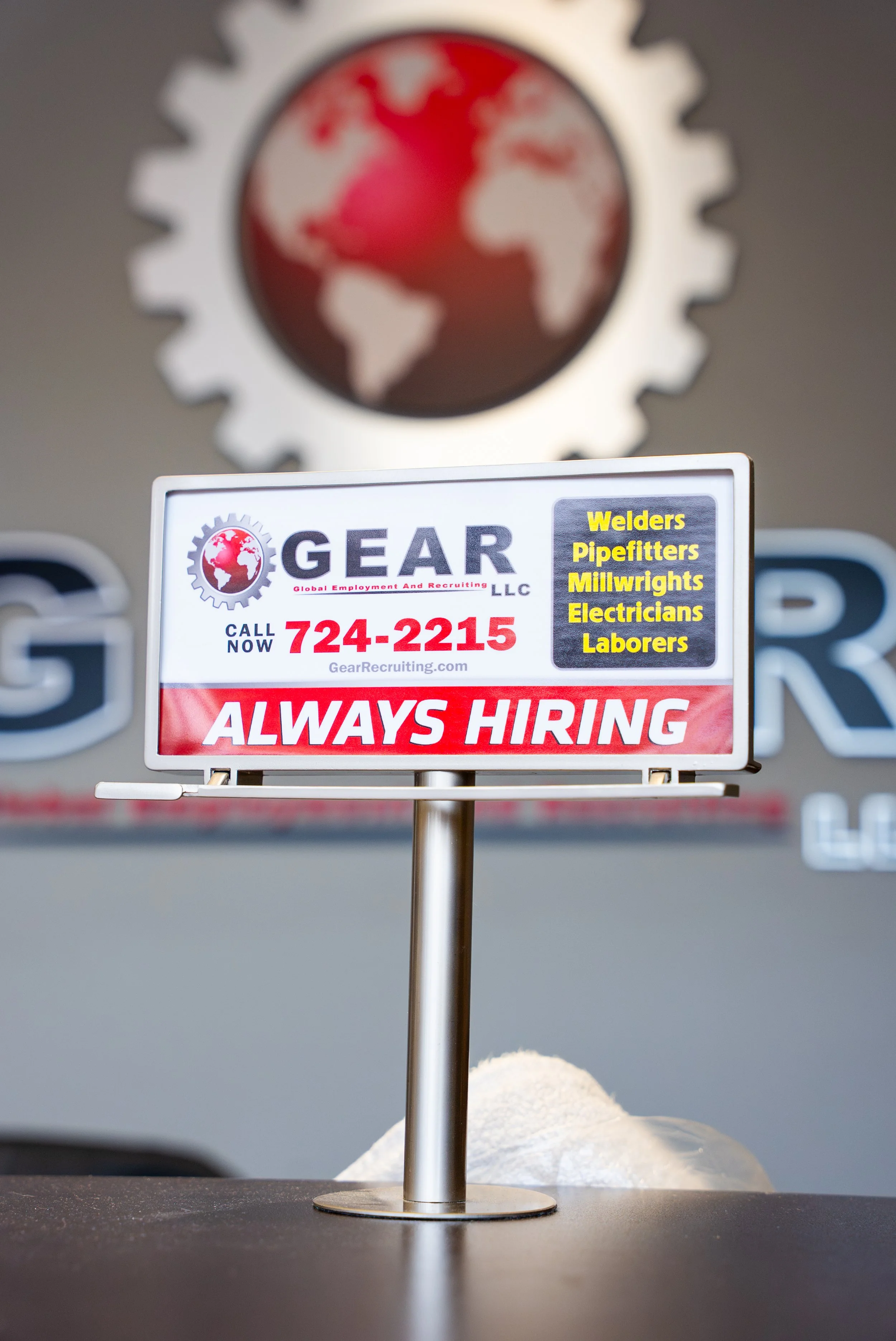 A sign with the GEAR LLC logo and contact information, listing trades such as welders, pipe fitters, millwrights, electricians, and laborers, with the words 'Always Hiring' in bold red.