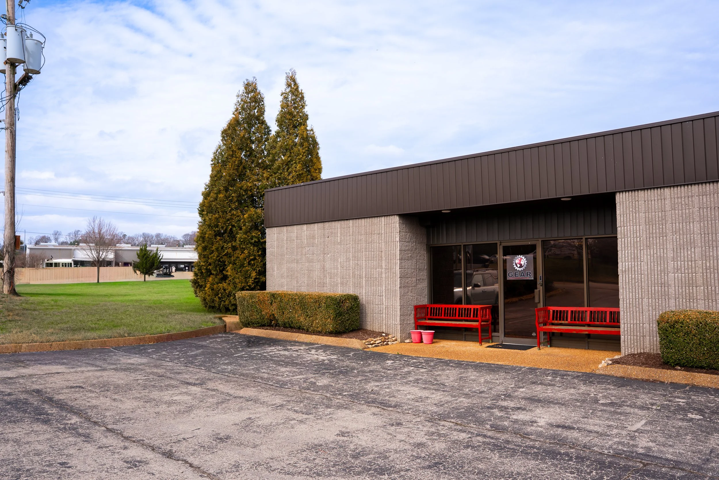 A small commercial building with a dark brown roof and a brick exterior wall, two red benches outside, and a parking lot in front. There are shrubs and trees around the building and a cloudy sky overhead.