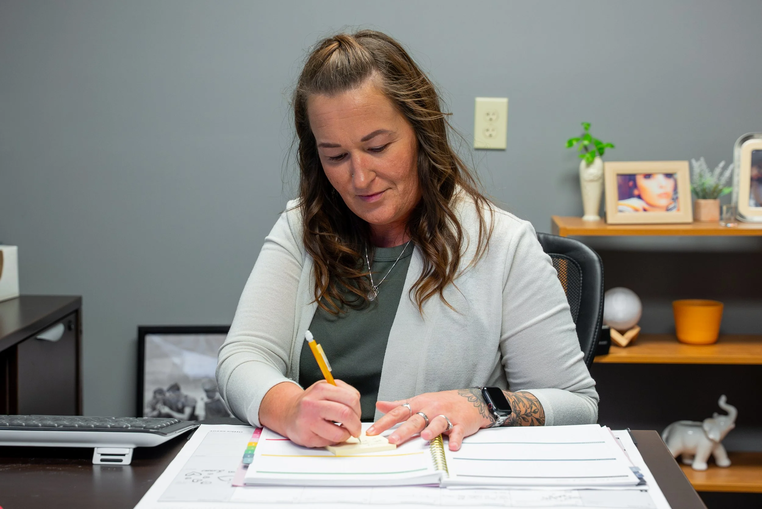 Woman sitting at a desk writing on a notepad with a pencil, in an office environment.