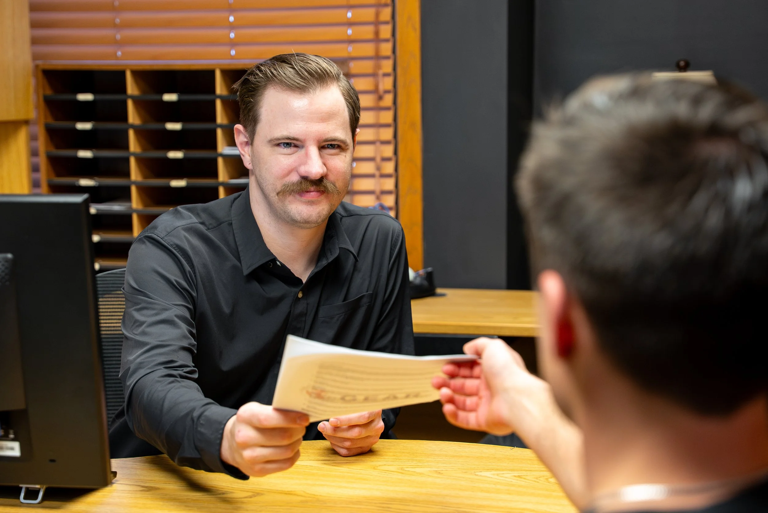 Man in black shirt handing a document to another person in an office.