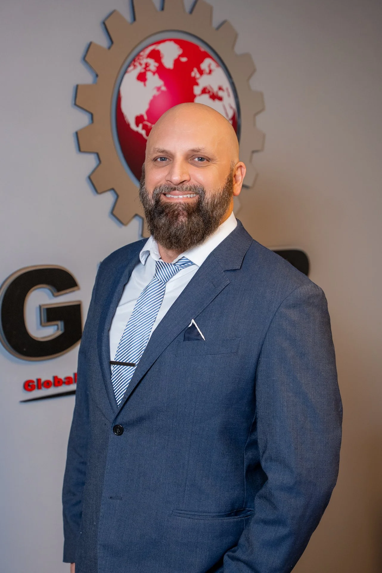 A man in a blue suit and striped tie standing in front of a logo with a globe and gear design. The background has the words 'Global' visible.