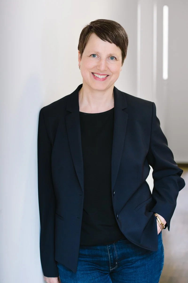 Eine lachende Frau mit kurzen braunen Haaren trägt eine dunkle Blazer und Jeans und steht in einem hellen Raum.