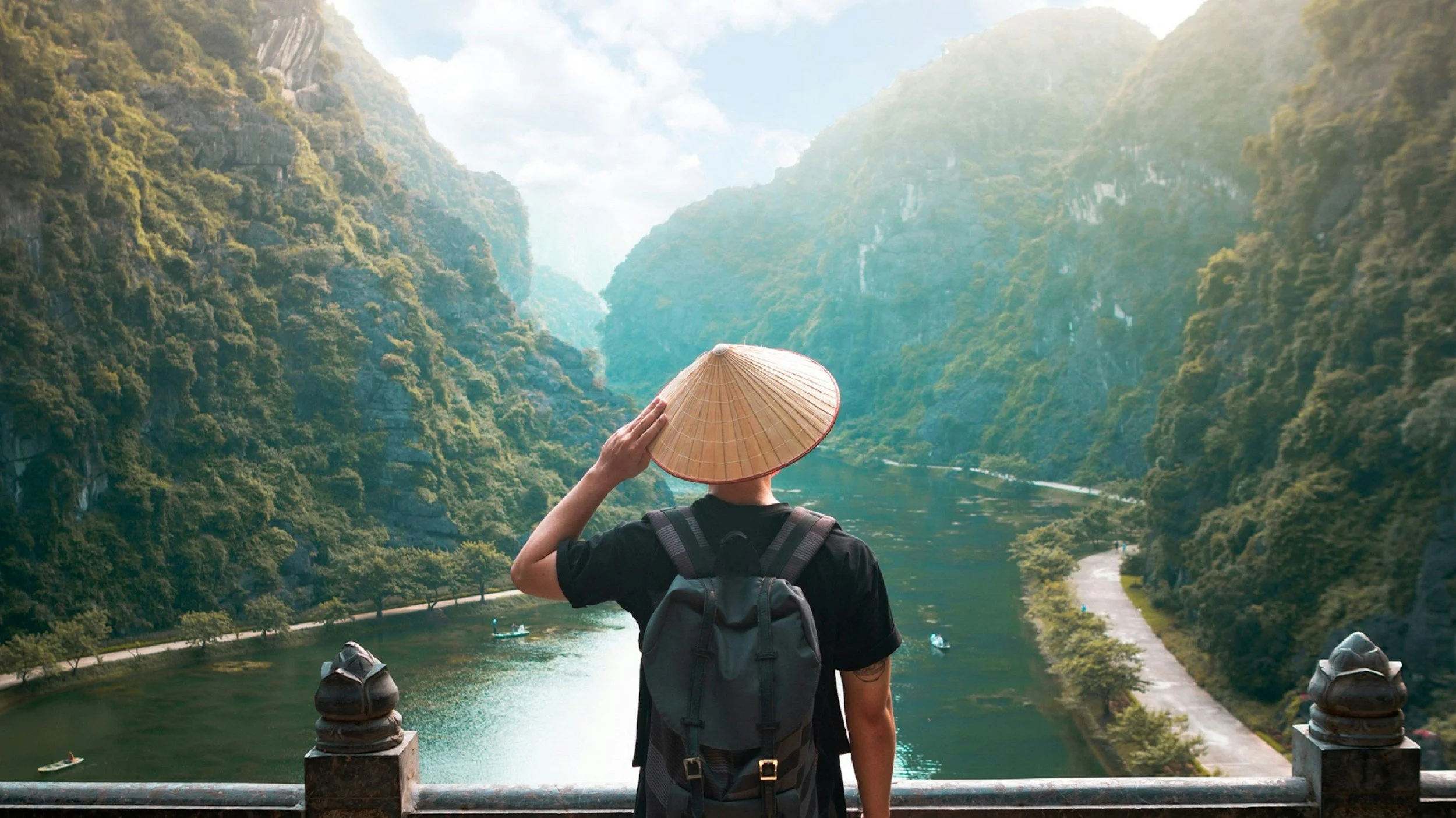 A person with a backpack holding a traditional Asian conical hat, overlooking a river with boats surrounded by lush green mountains.