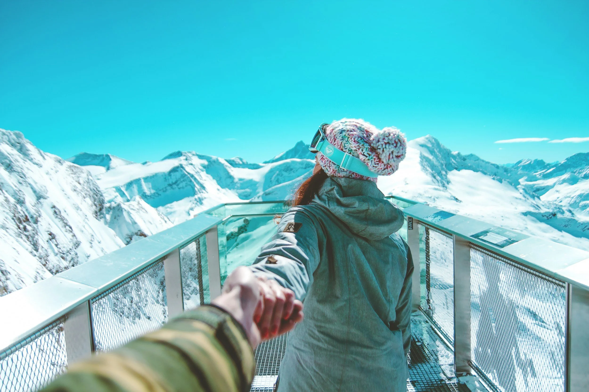 Person holding hands with another person on a glass platform with snow-covered mountain peaks and blue sky in the background