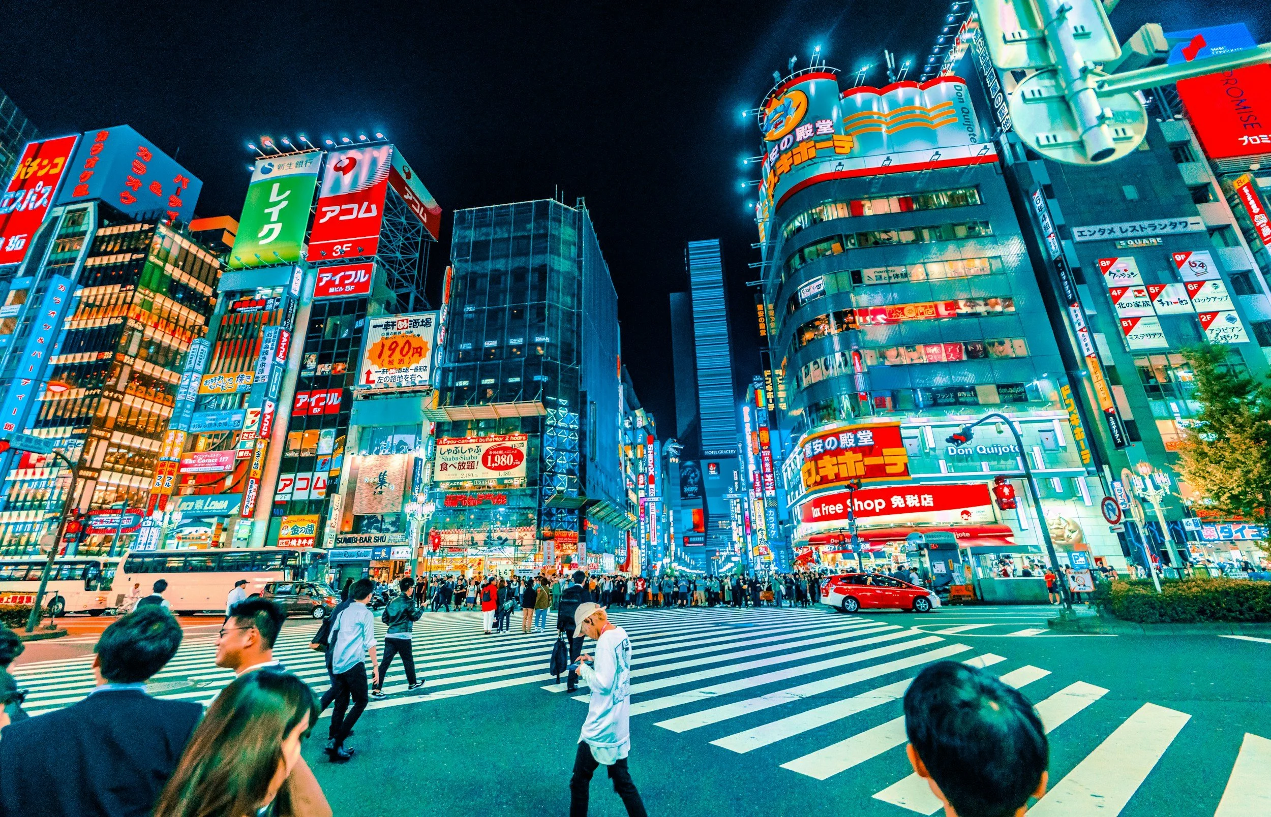 Nighttime city scene in Tokyo with brightly lit buildings, colorful advertising signs, and a busy crosswalk filled with pedestrians.