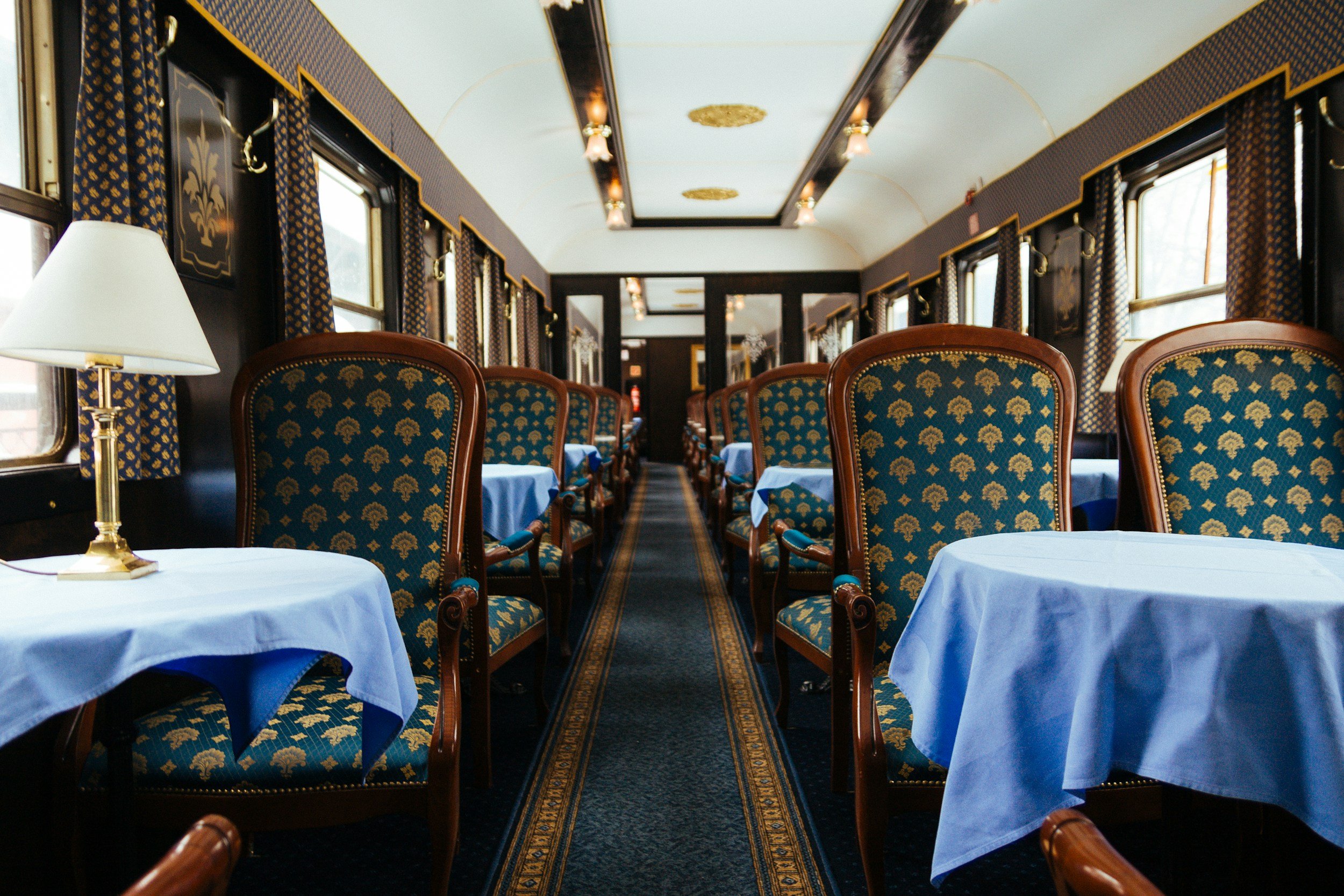 Elegant dining room inside a vintage train with round tables covered with blue tablecloths, wooden chairs with patterned upholstery, lamps, and large windows with curtains.