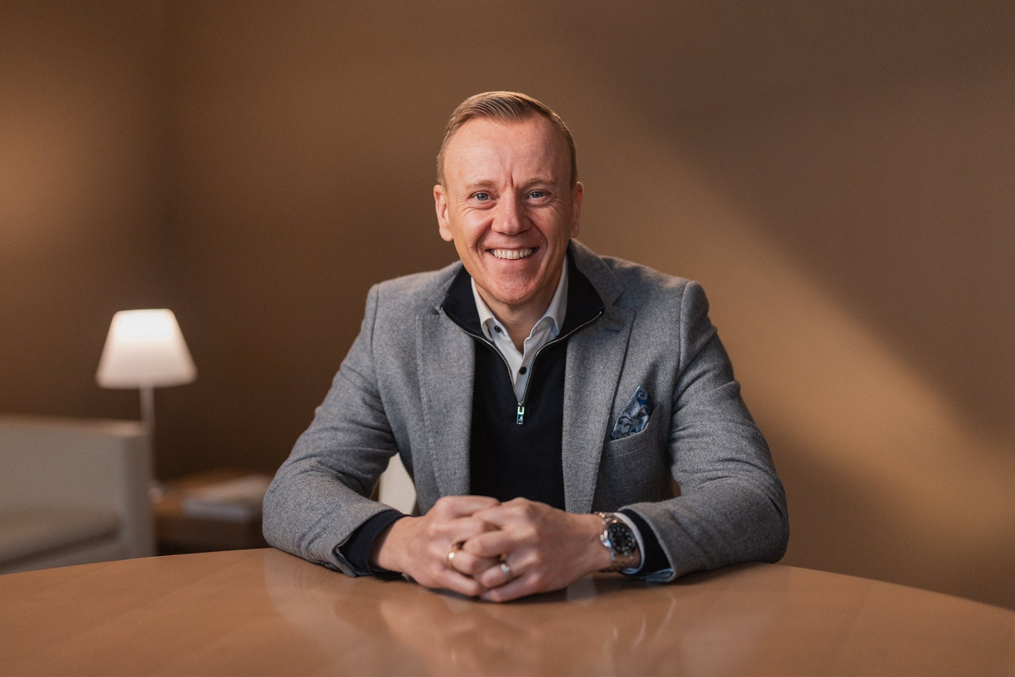 A smiling man with red hair, dressed in a grey blazer and black shirt, sitting at a table in a warmly lit room.