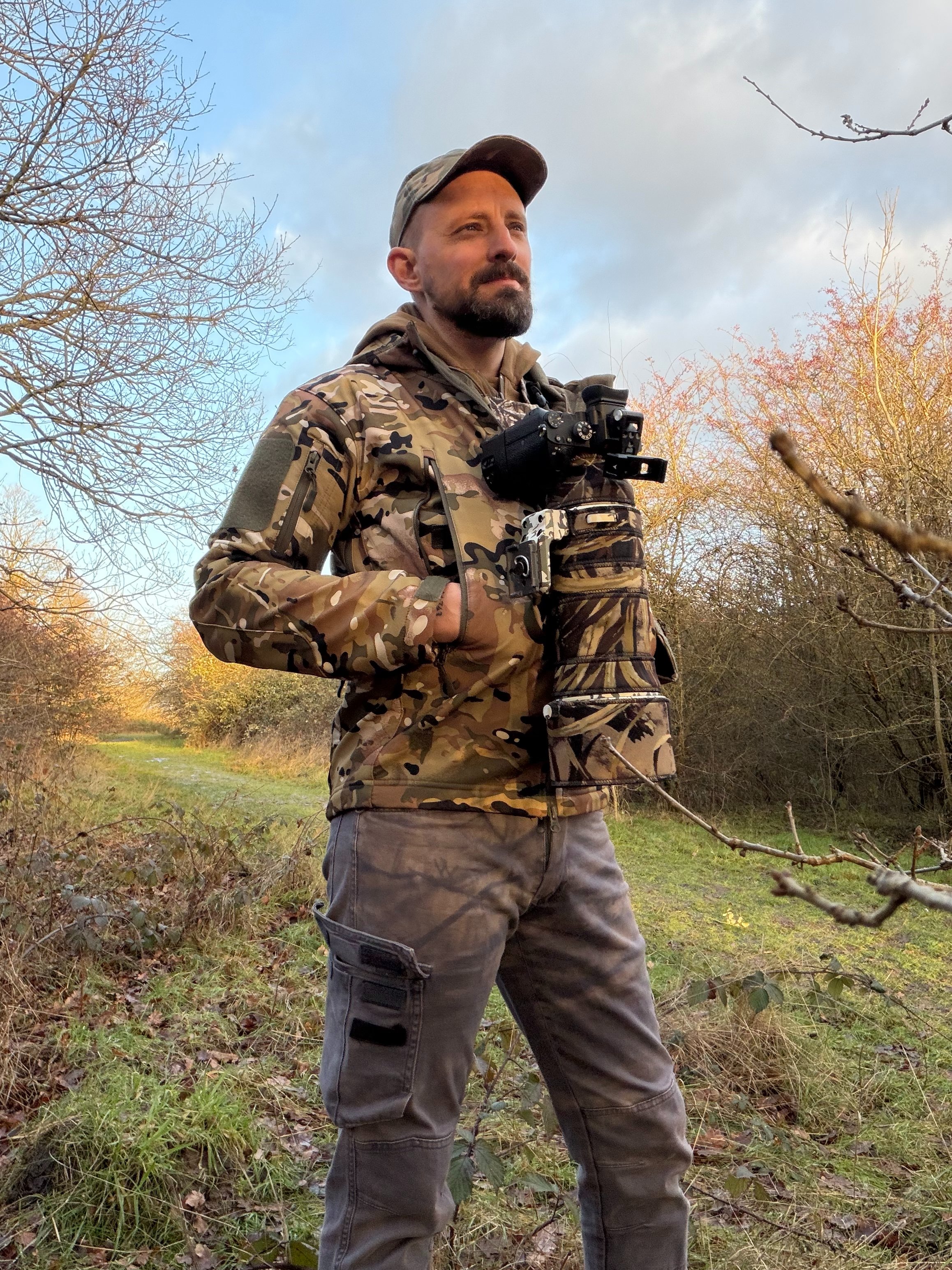A man in camouflage outdoors holding a camera with a jungle camouflage cover, standing on a grassy trail with trees and shrubs around on a partly cloudy day.