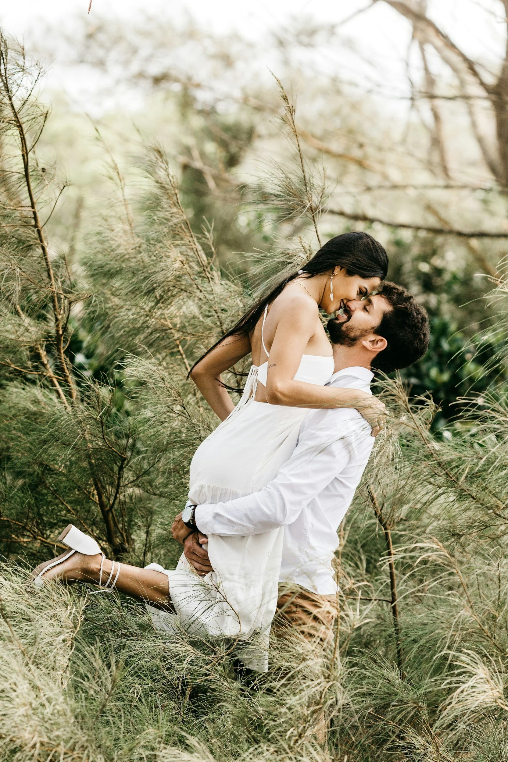 A couple in white clothing embracing in a natural outdoor setting among tall grass and trees, with the woman lifted by the man.