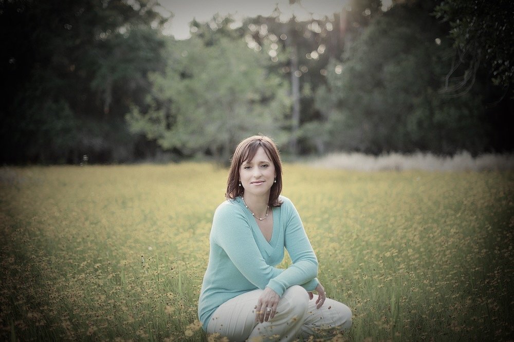 A woman sitting cross-legged on the ground in a field of grass and yellow flowers, surrounded by trees, during sunset, looking at the camera.