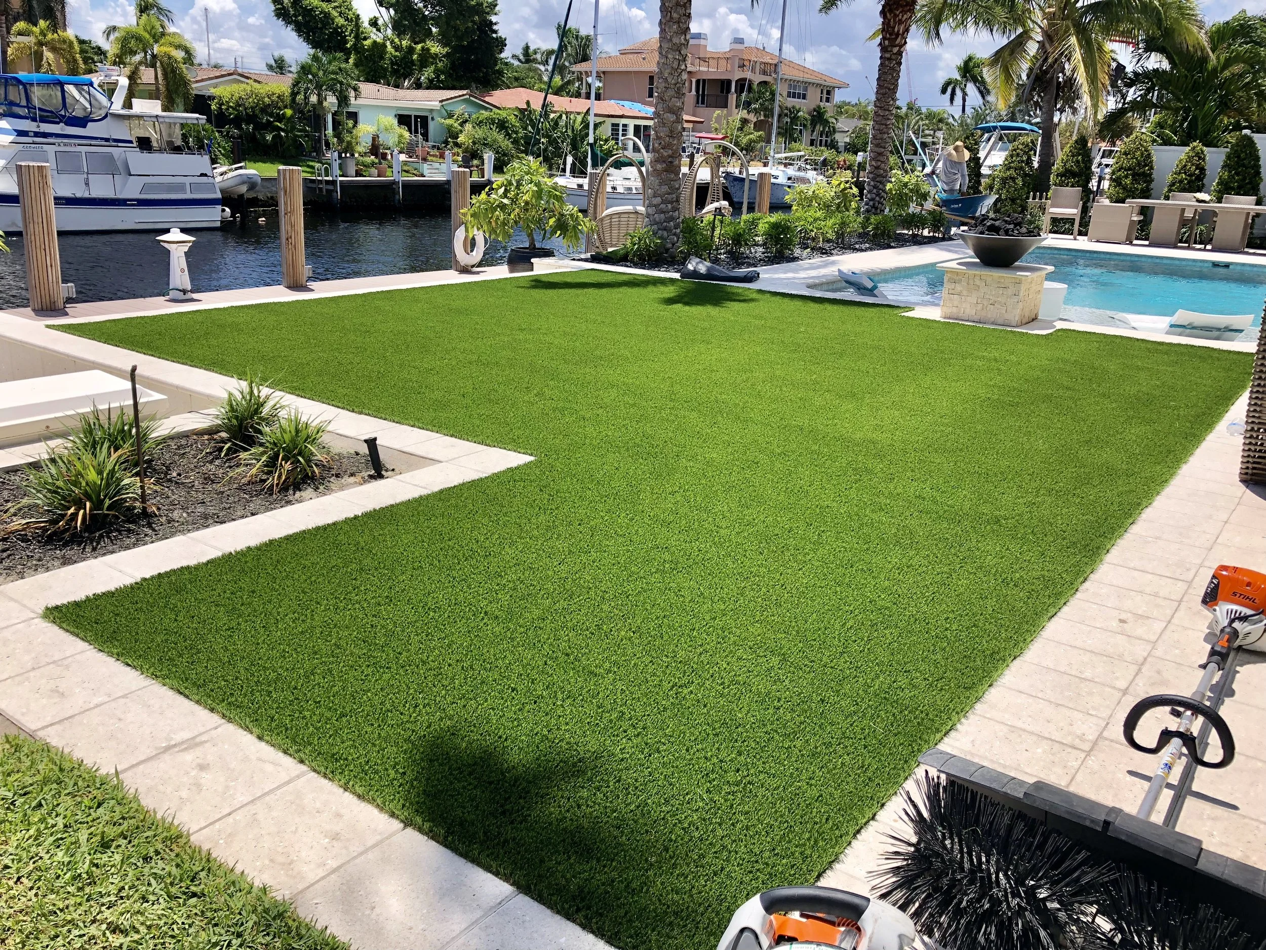 Backyard with lush artificial grass, next to a paved patio area, overlooking a canal with boats and palm trees, with a swimming pool and outdoor seating in the background.