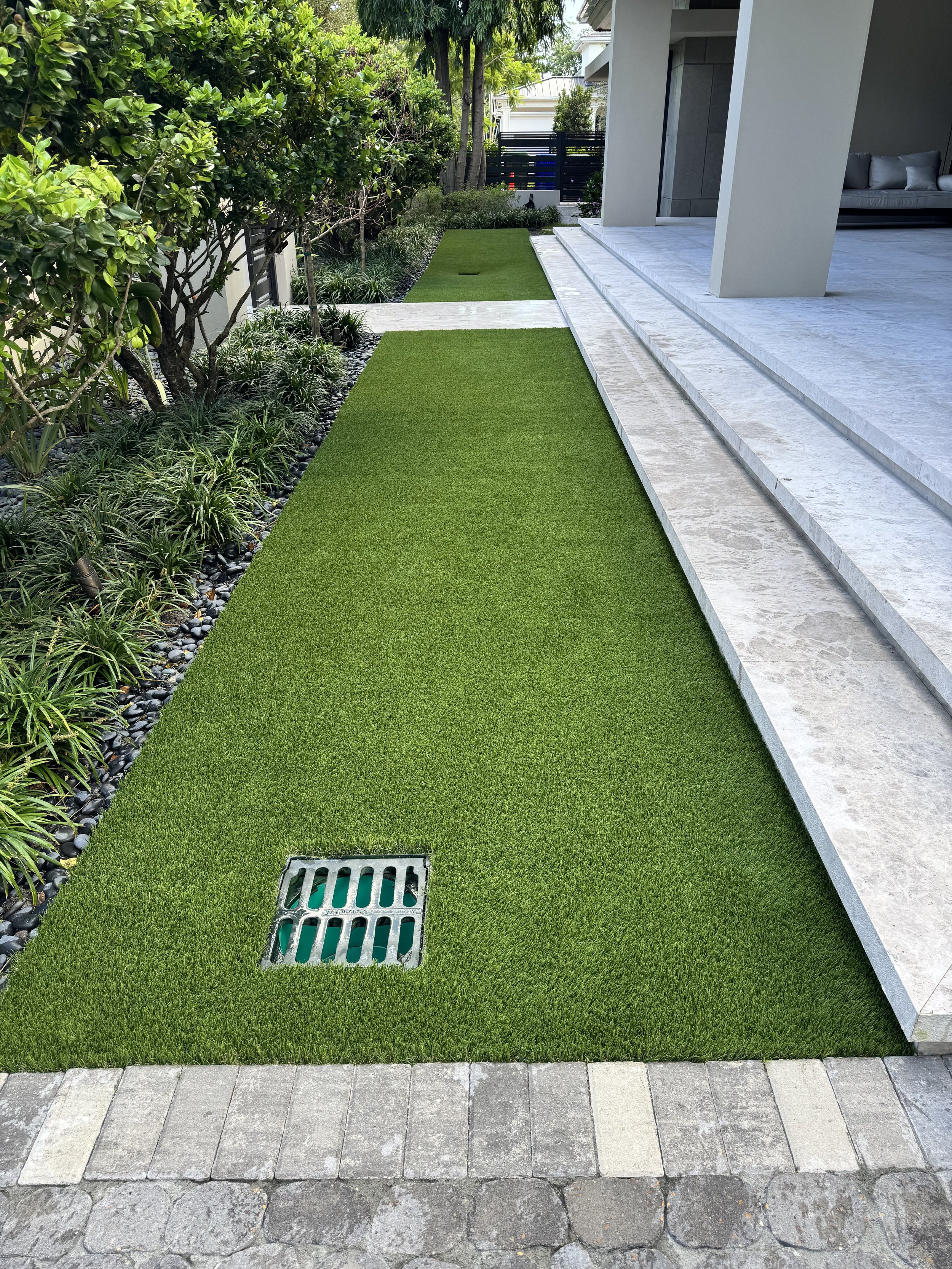 Artificial grass lawn bordered by small decorative rocks and enclosed by bushes and trees alongside a modern building with a stone patio and steps.