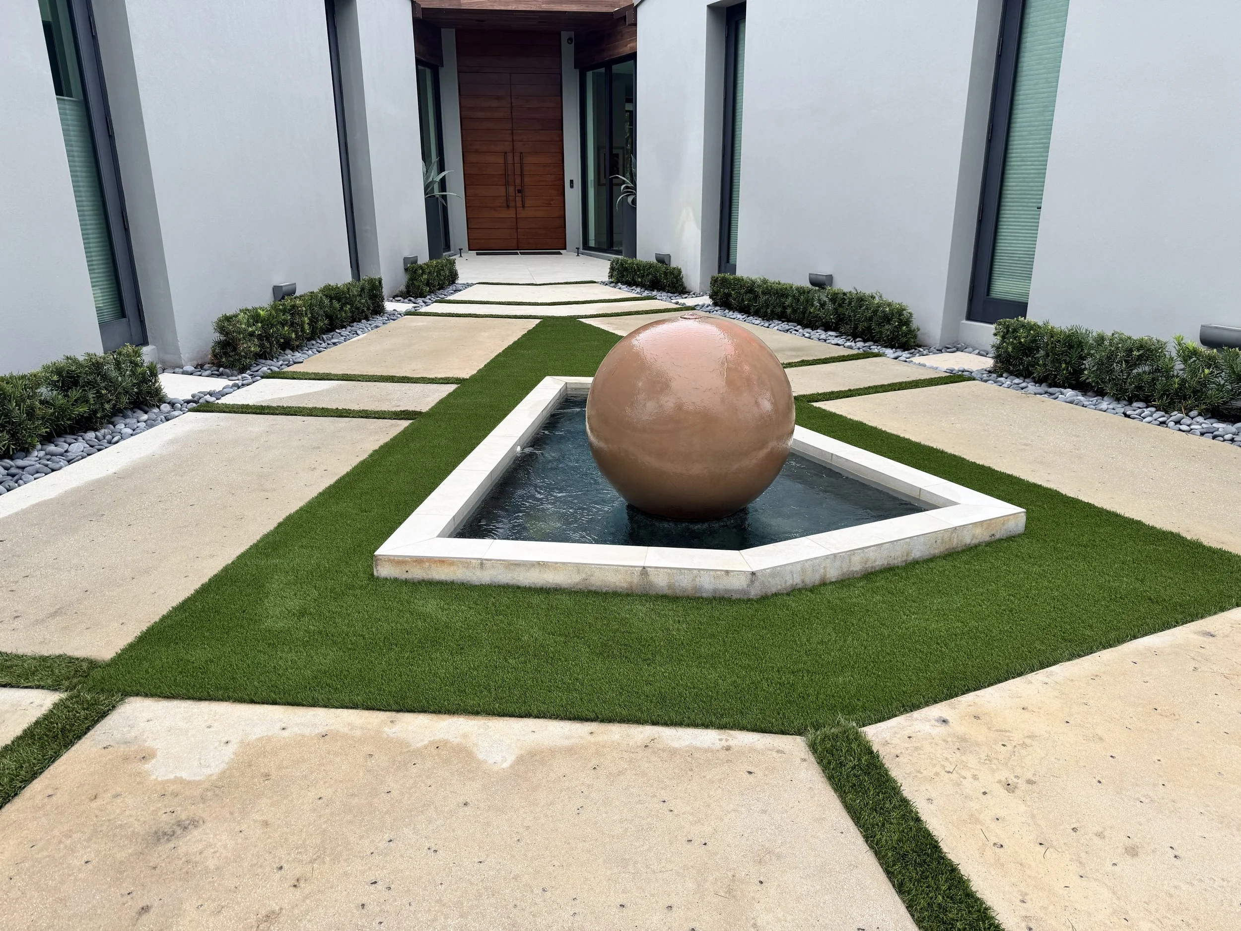 Modern front yard with a small rectangular fountain featuring a large spherical sculpture, surrounded by well-kept green grass, concrete pathways, and concrete edging. The house has a large wooden front door, light-colored exterior walls, and tall windows.