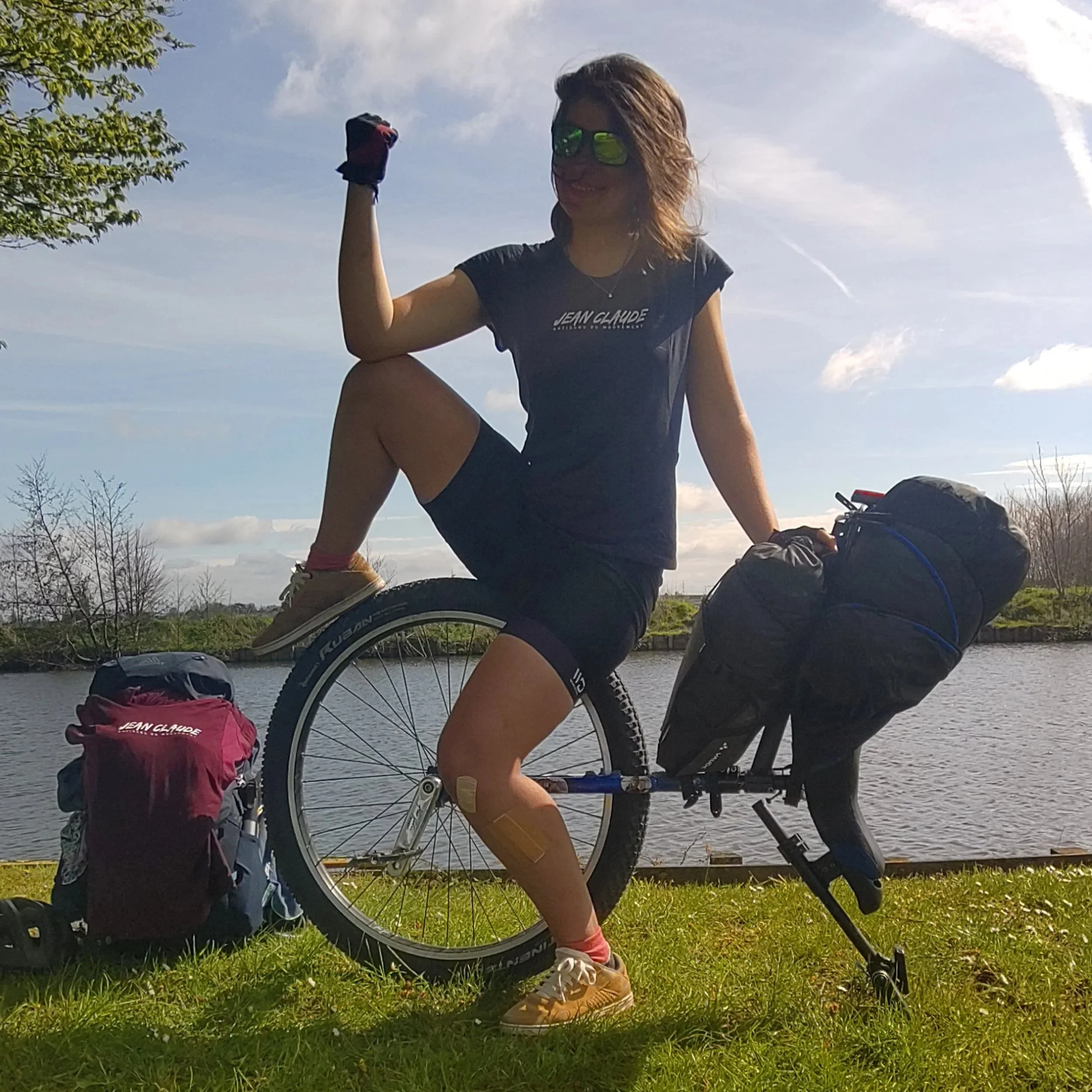Femme assise sur un monocycle, faisant un geste de victoire, portant des lunettes de soleil, avec un lac en arrière-plan.