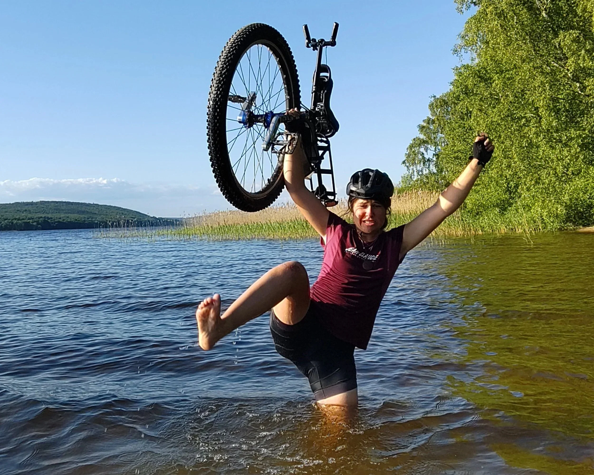 Une femme souriante, portant un casque de vélo, se tient dans l'eau avec une jambe levée et un bras levé, tenant un monocycle au-dessus de sa tête, dans un environnement naturel avec de l'eau, des arbres et un ciel clair.