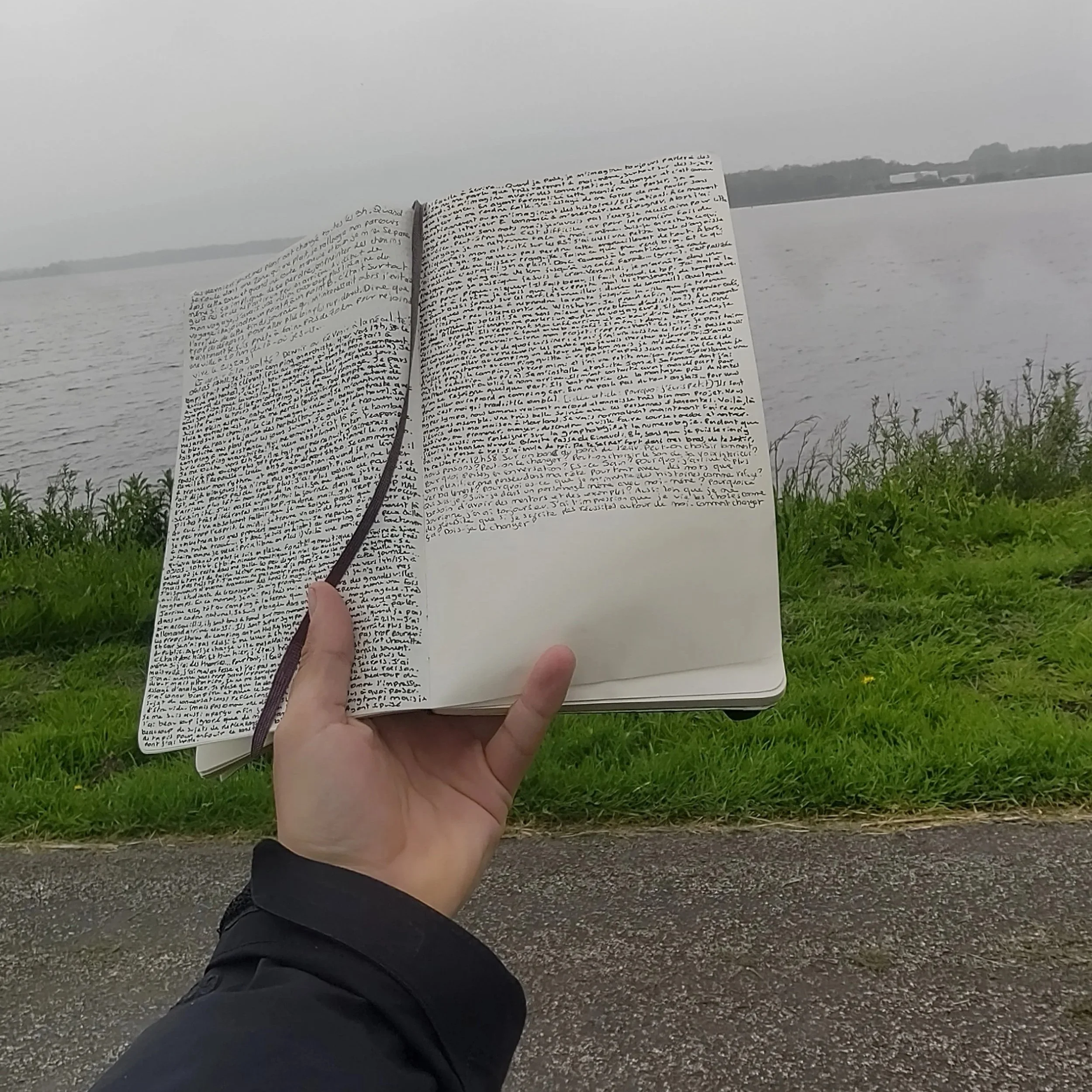 Une main tient un carnet ouvert avec beaucoup d'écriture manuscrite proche d'un lac ou d'une rivière par une journée nuageuse.