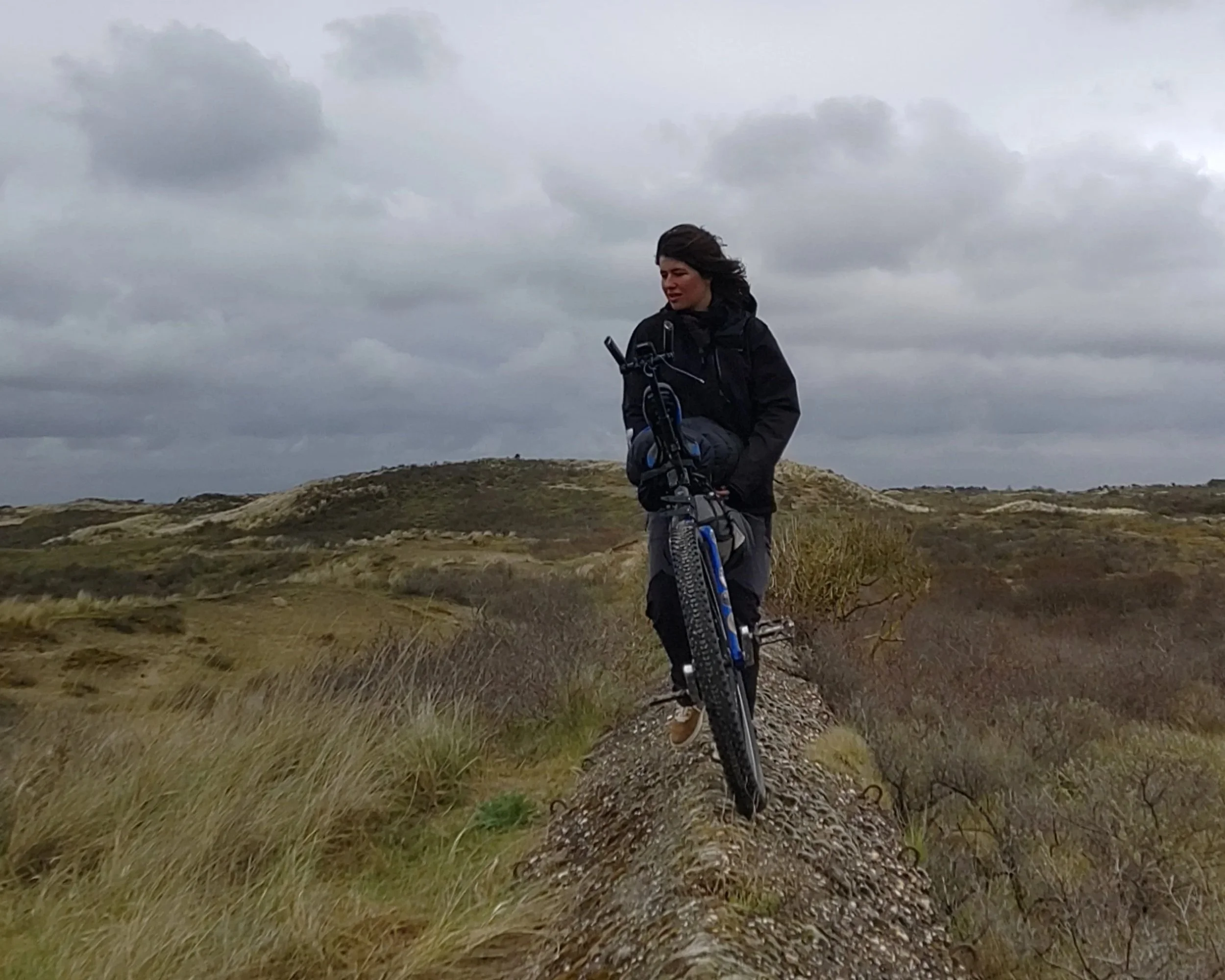 Une femme en vêtements noirs son monocycle à la main, avance sur une petite crête dans un paysage de collines sous un ciel nuageux.